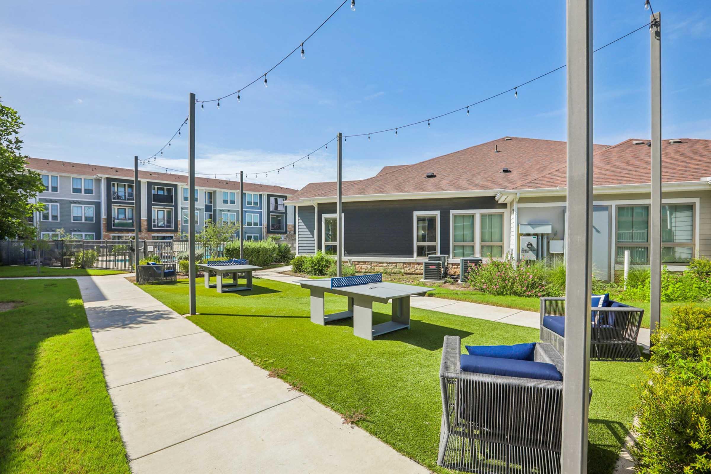 A landscaped outdoor lounge area featuring green grass, string lights overhead, and modern furniture including tables and chairs. In the background, a residential building is visible. The setting is bright and inviting, suitable for relaxation and socializing.