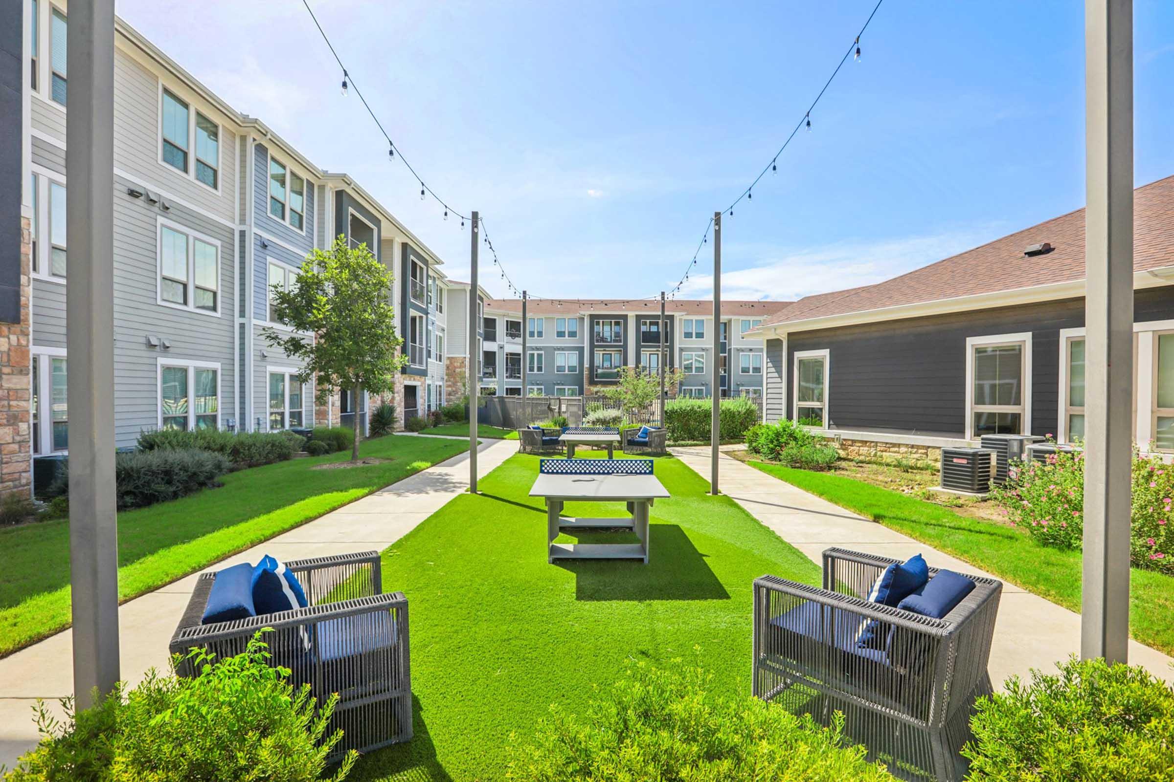 A landscaped courtyard area featuring green artificial grass, lounge chairs, and a ping pong table, surrounded by modern apartment buildings. String lights hang above, adding a cozy ambiance to the outdoor space, perfect for relaxation and socializing.