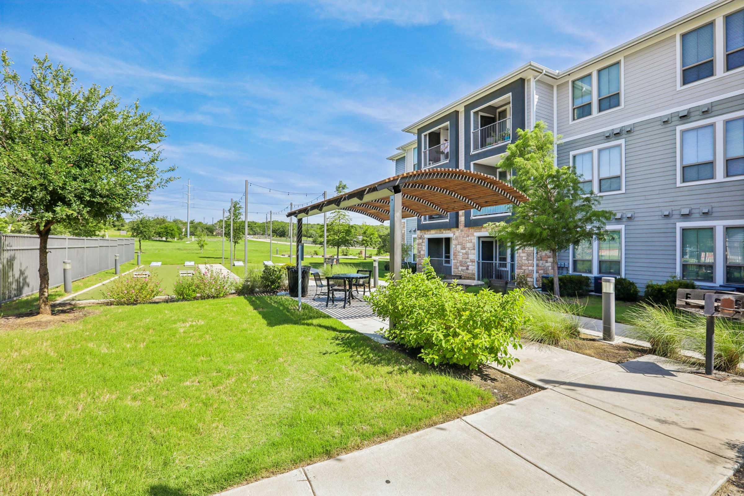 A landscaped area featuring a wooden pergola with seating, surrounded by grass and trees. In the background, there are modern apartment buildings and a green space, creating a pleasant outdoor environment. Clear blue skies enhance the inviting atmosphere.