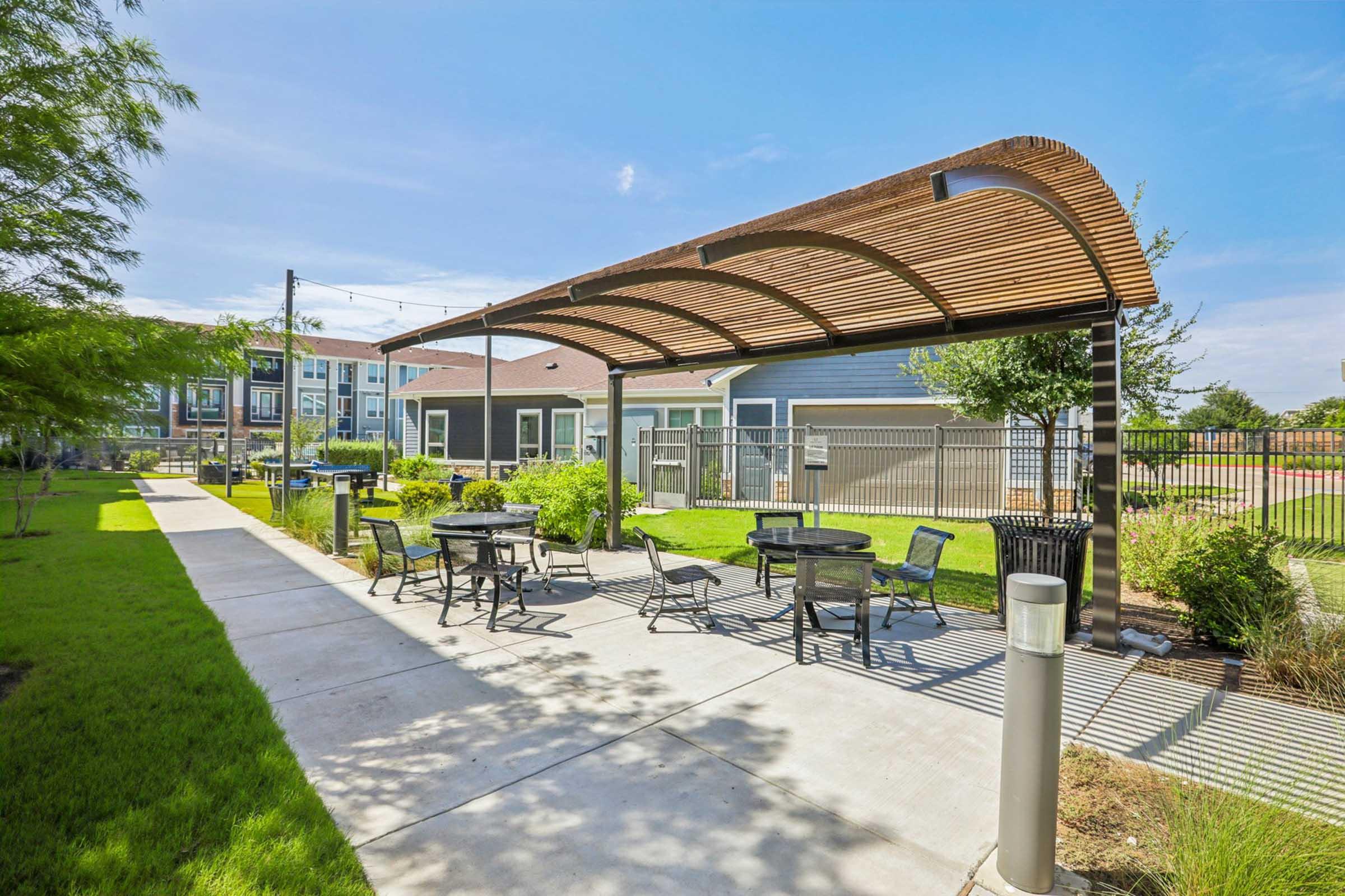 Outdoor area featuring a paved walkway lined with green grass, shaded seating under a wooden structure, and modern buildings in the background. The scene is bright and sunny, showcasing a well-maintained landscaped environment.