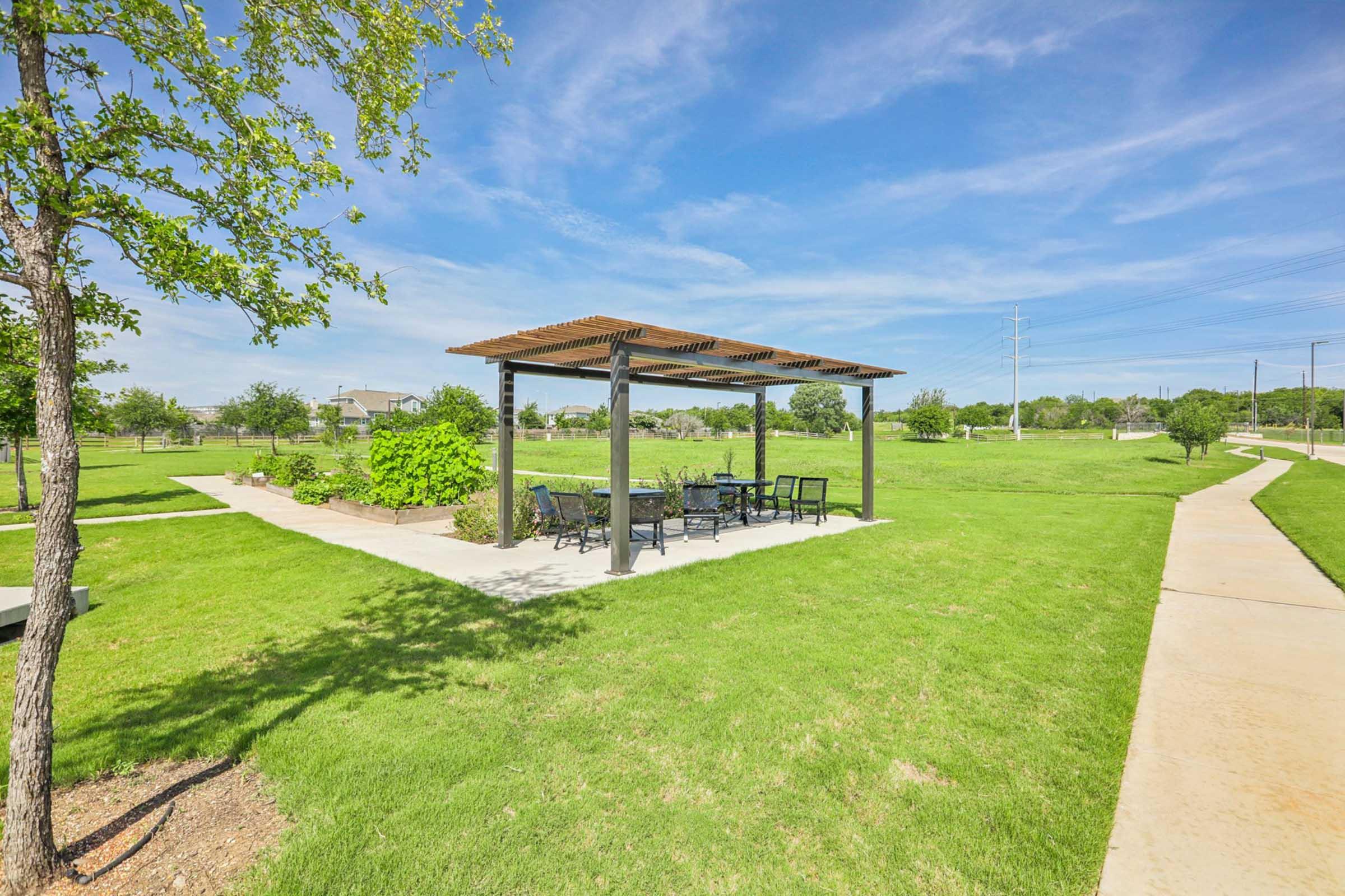 A sunny park scene featuring a shaded gazebo with a picnic table and chairs, surrounded by green grass and trees. A paved pathway runs alongside the area, leading into the open space with blue skies and scattered clouds overhead.
