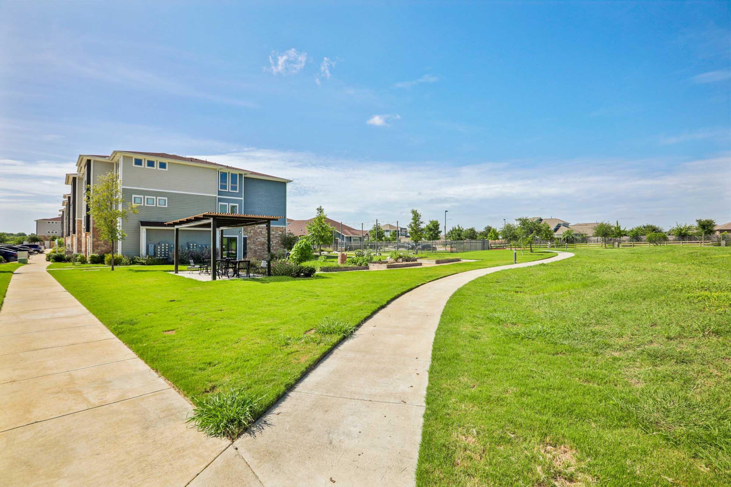 A pathway winds through a landscaped area with green grass and trees, leading to a multi-story building. In the foreground, there are benches and recreational spaces, with an open sky above. The scene conveys a peaceful and inviting outdoor environment in a residential area.