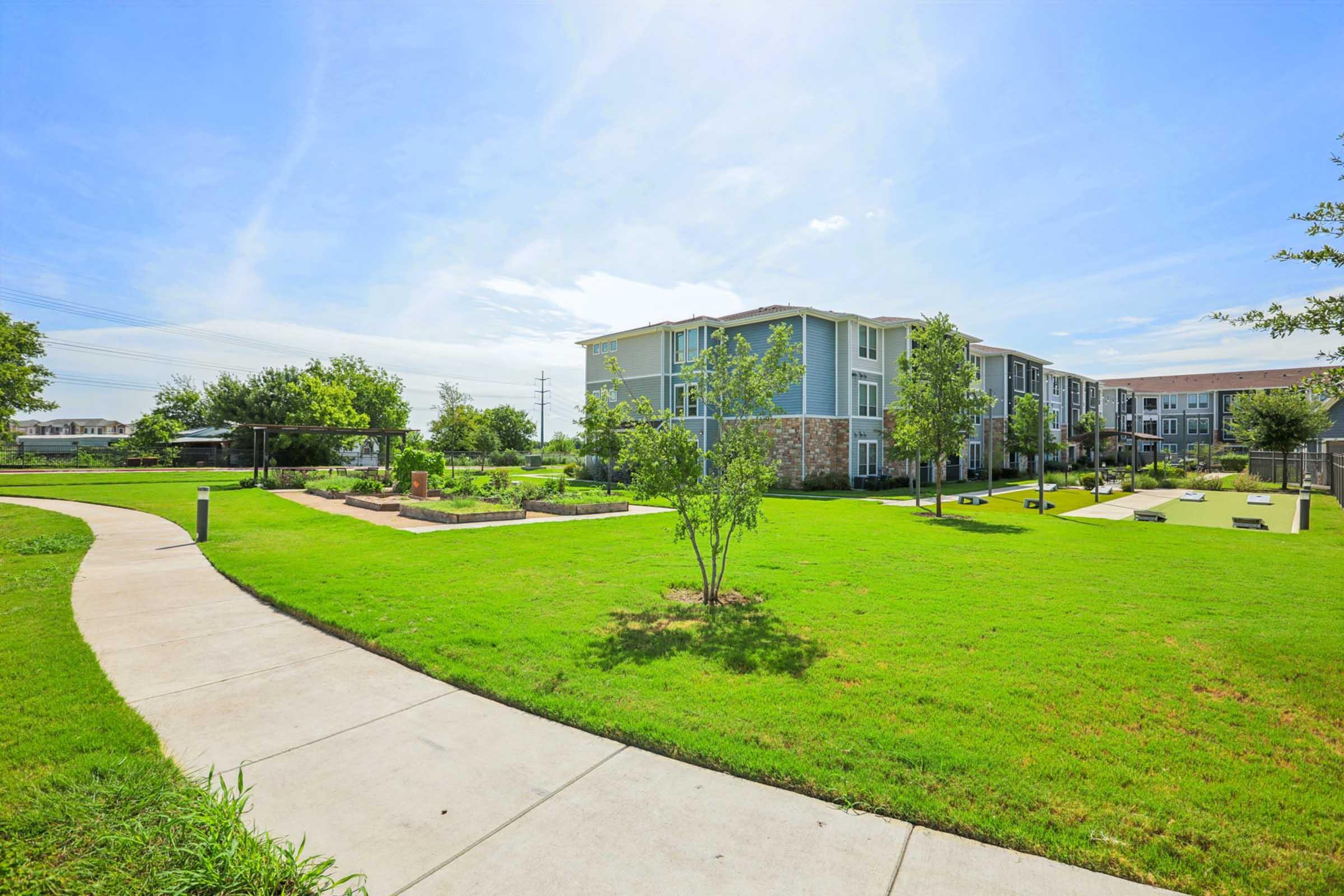 A bright, landscaped area featuring a paved walking path, lush green grass, and young trees. In the background, there are modern apartment buildings. The sky is clear and blue, suggesting a sunny day. The scene conveys a peaceful, inviting outdoor space.