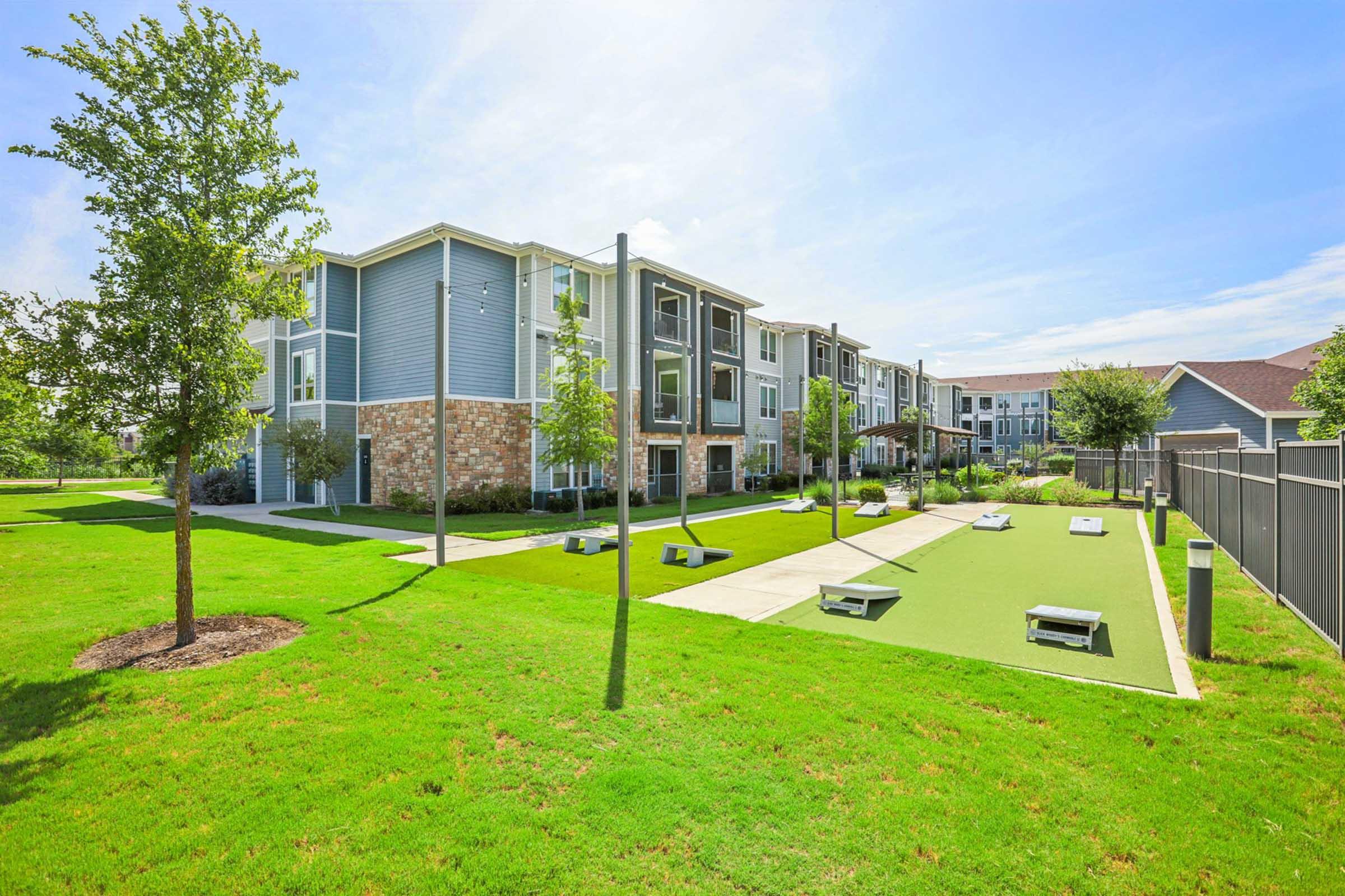A view of a modern apartment complex surrounded by well-maintained green lawns and trees. The area features outdoor seating and a grassy activity space, with clear blue skies overhead. The buildings exhibit a blend of brick and siding, contributing to an inviting residential atmosphere.