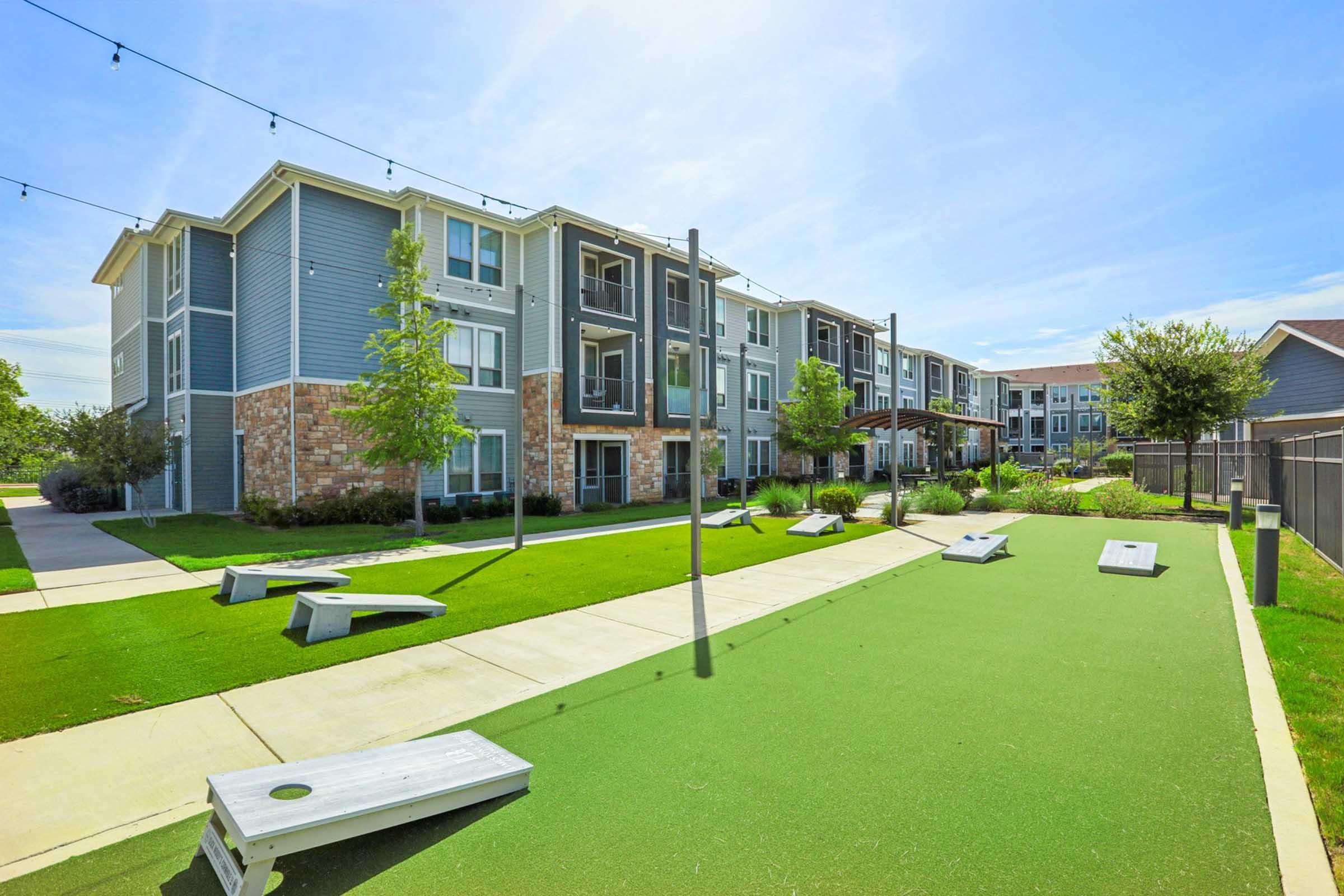 A well-maintained outdoor area featuring multi-story apartment buildings with balconies, surrounded by greenery. There are several cornhole game boards set up on a bright green turf area, with clear skies above and string lights adding ambiance.