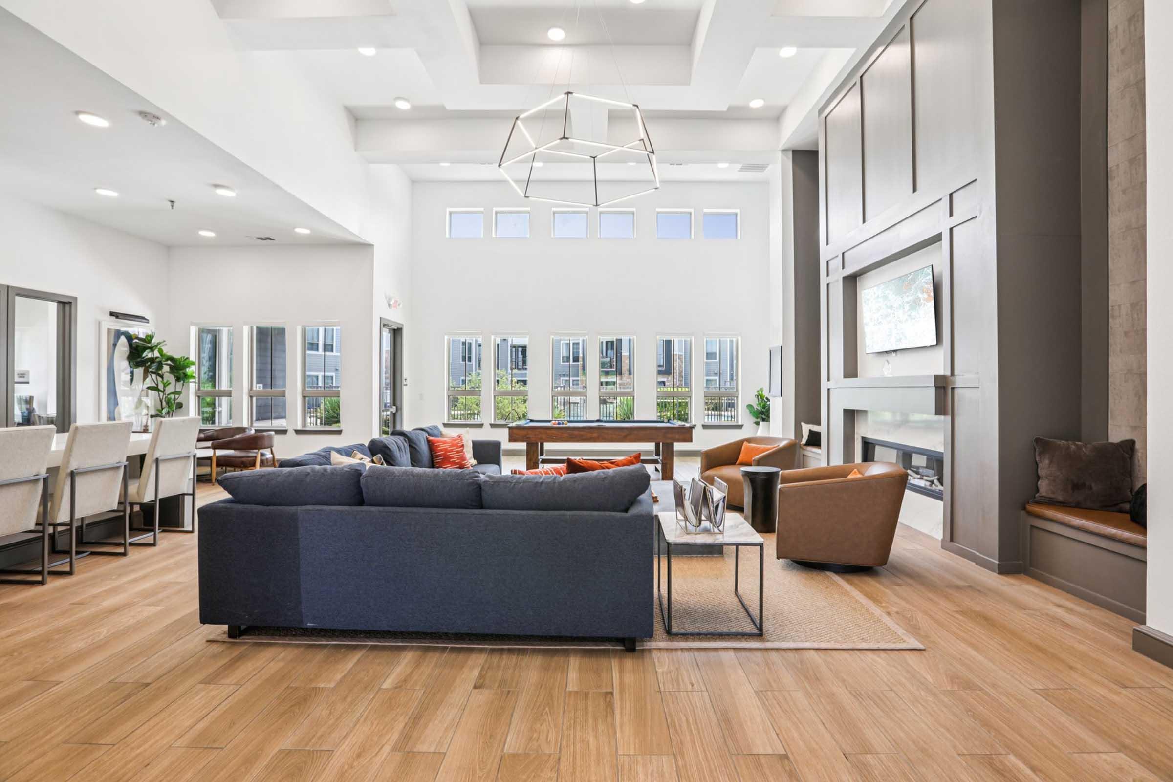 Spacious modern living area featuring a comfortable blue sectional sofa, brown armchairs, and a coffee table. Large windows allow natural light, complemented by a stylish chandelier overhead. A sleek fireplace and contemporary decor create an inviting atmosphere.
