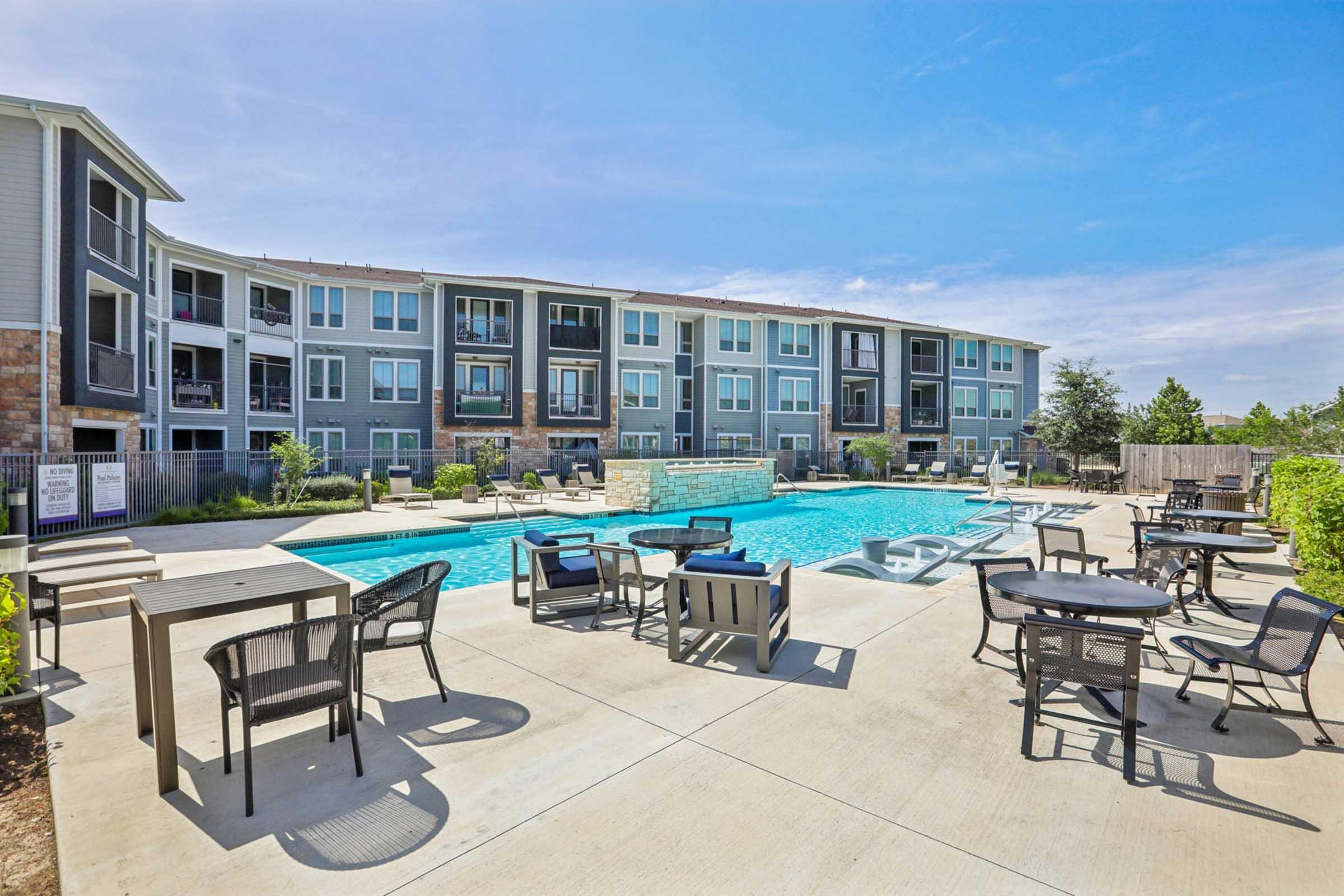 A bright outdoor pool area featuring a large swimming pool surrounded by lounge chairs and tables. The backdrop consists of a stylish multi-story apartment building with large windows, under a clear blue sky. Lush greenery and fences enhance the relaxing atmosphere of the space.