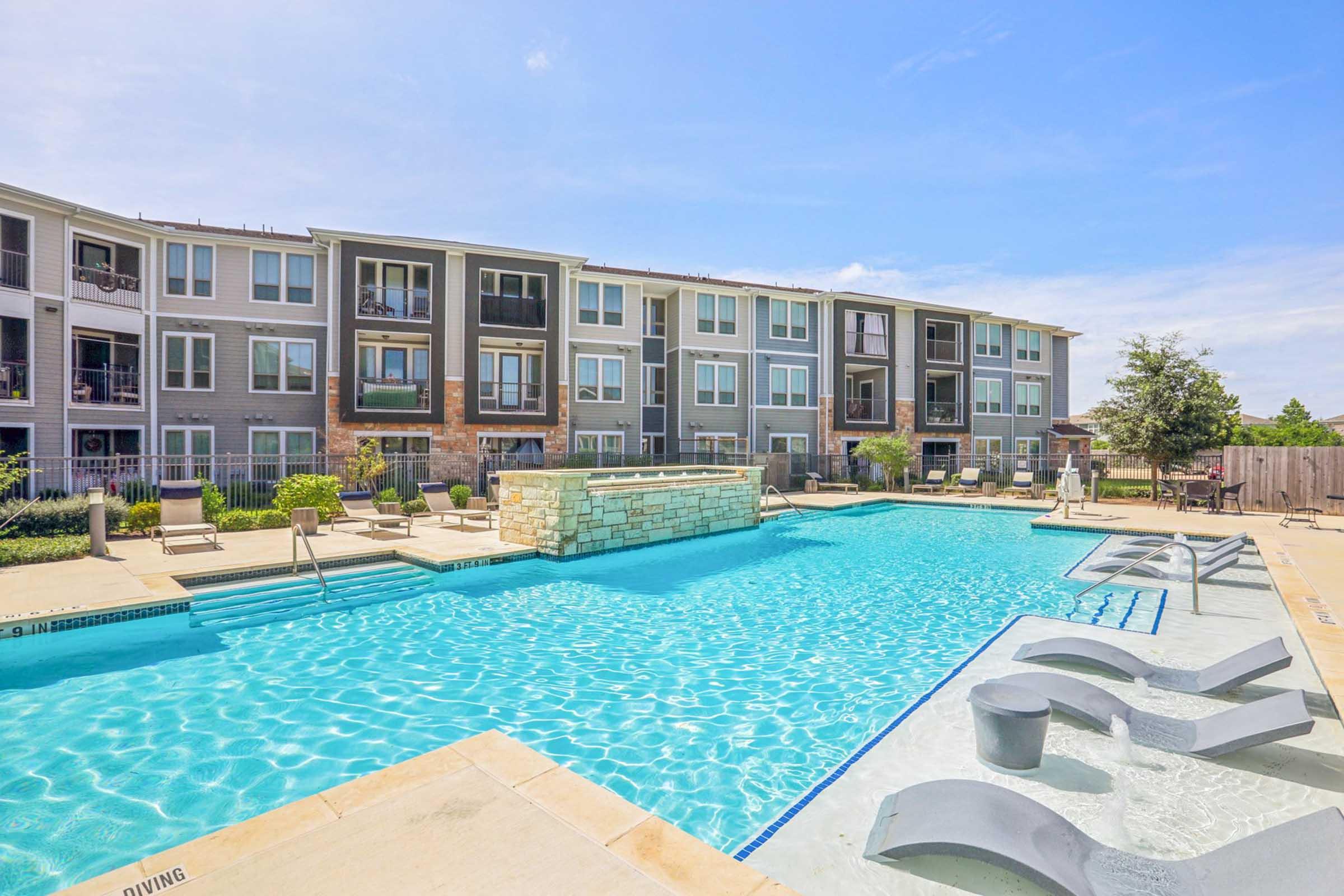 A clear blue swimming pool surrounded by well-maintained lounge chairs and landscaped areas. In the background, there are modern apartment buildings with multiple windows. The scene is bright and sunny, creating a relaxing atmosphere.