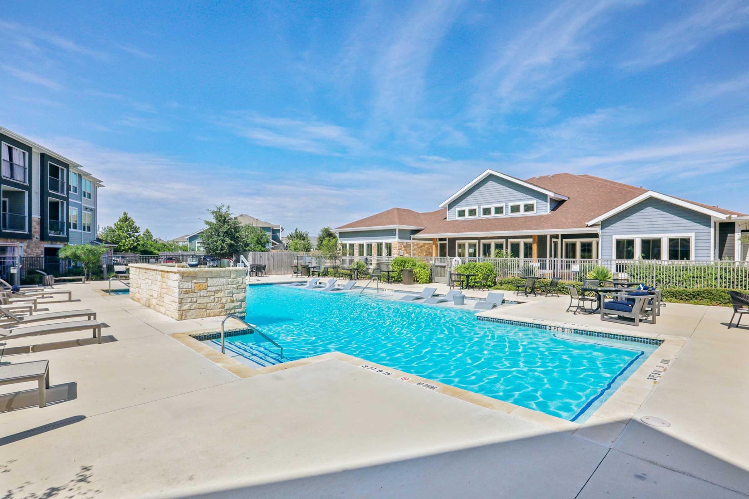A bright outdoor swimming pool with clear blue water, surrounded by lounge chairs. In the background, there’s a modern building with large windows and a sloped roof. The area is landscaped with trees and plants, under a clear blue sky, creating a relaxing atmosphere.