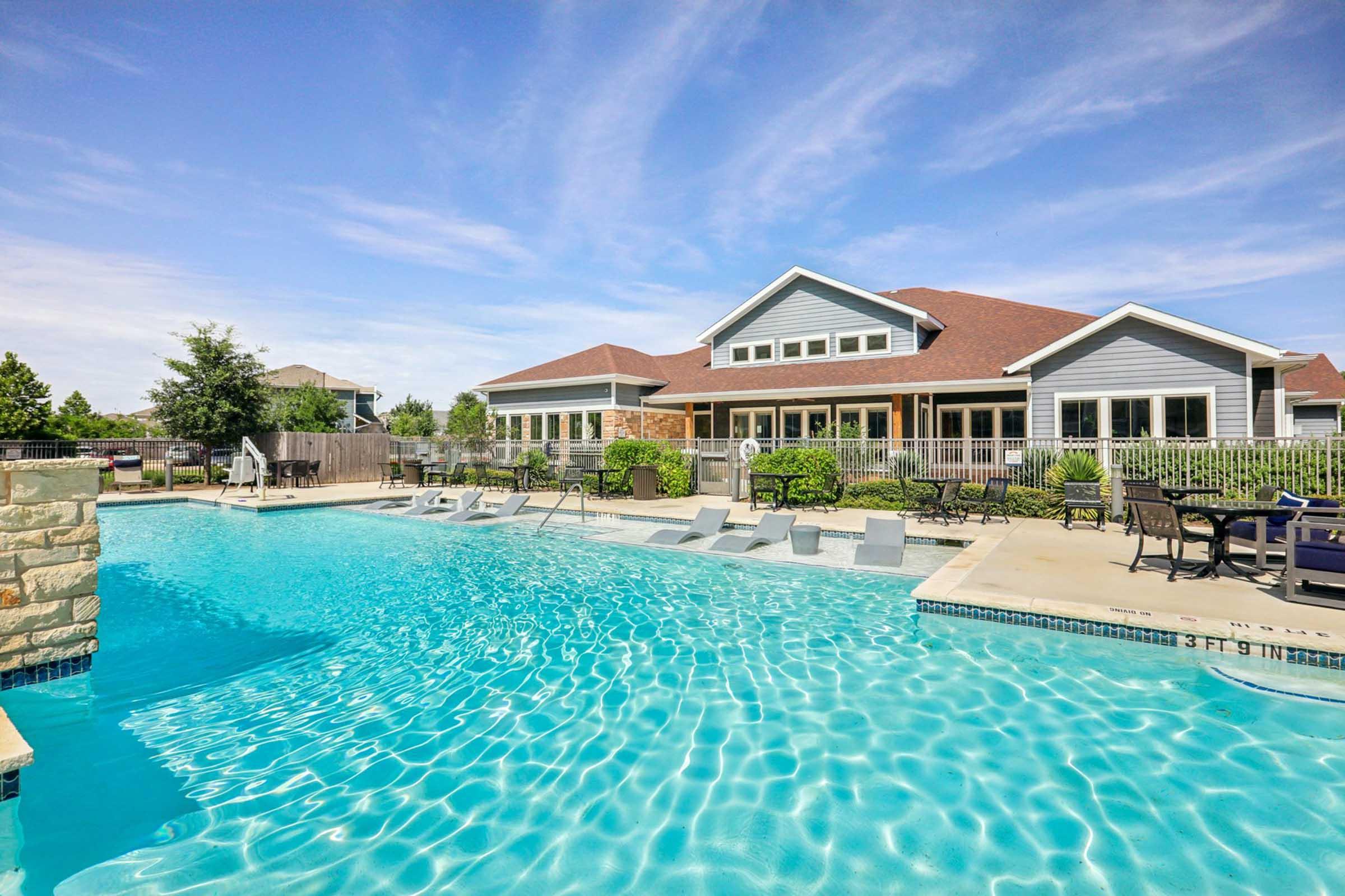 A sunny outdoor swimming pool surrounded by lounge chairs, with a modern building in the background featuring large windows and a sloped roof. Lush greenery and patio furniture add to the inviting atmosphere, making it an ideal spot for relaxation and leisure.
