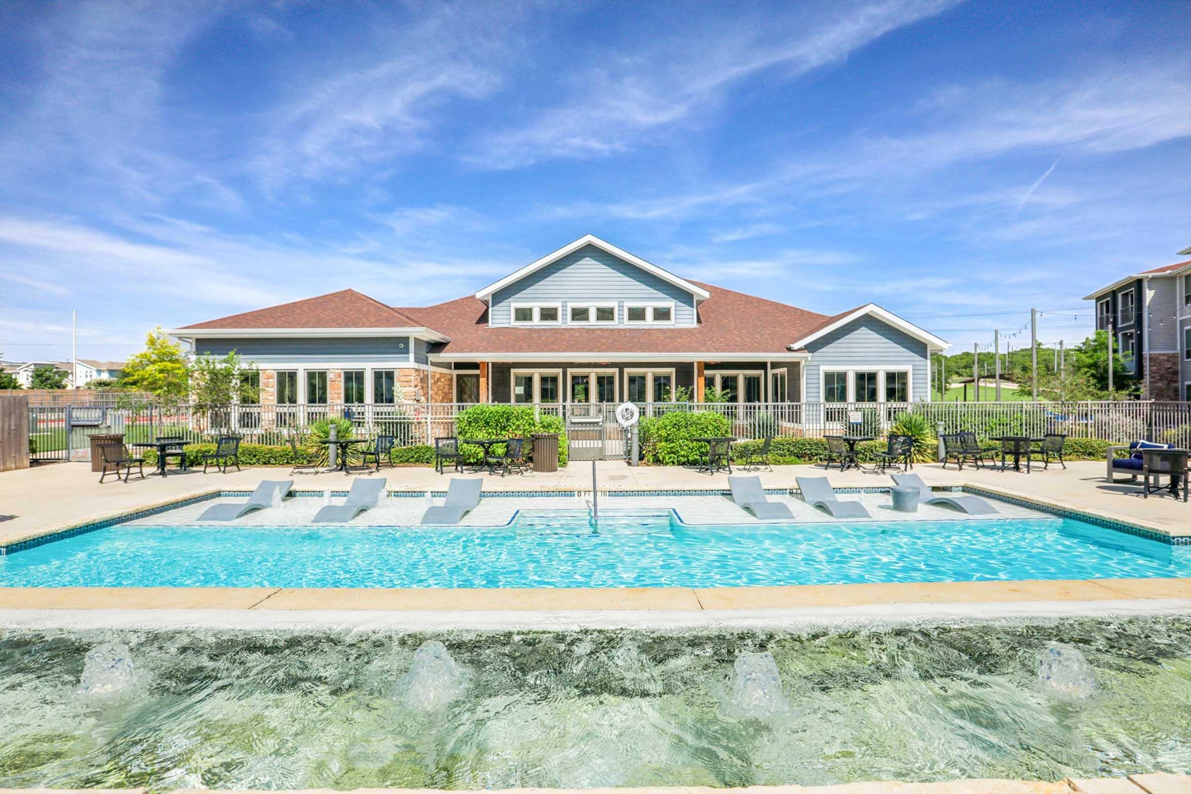 A modern clubhouse with a large swimming pool in front. The pool features lounge chairs along the edges and is surrounded by green landscaping. The building has large windows and a welcoming design, set against a clear blue sky with a few clouds. It's an inviting outdoor recreation area.