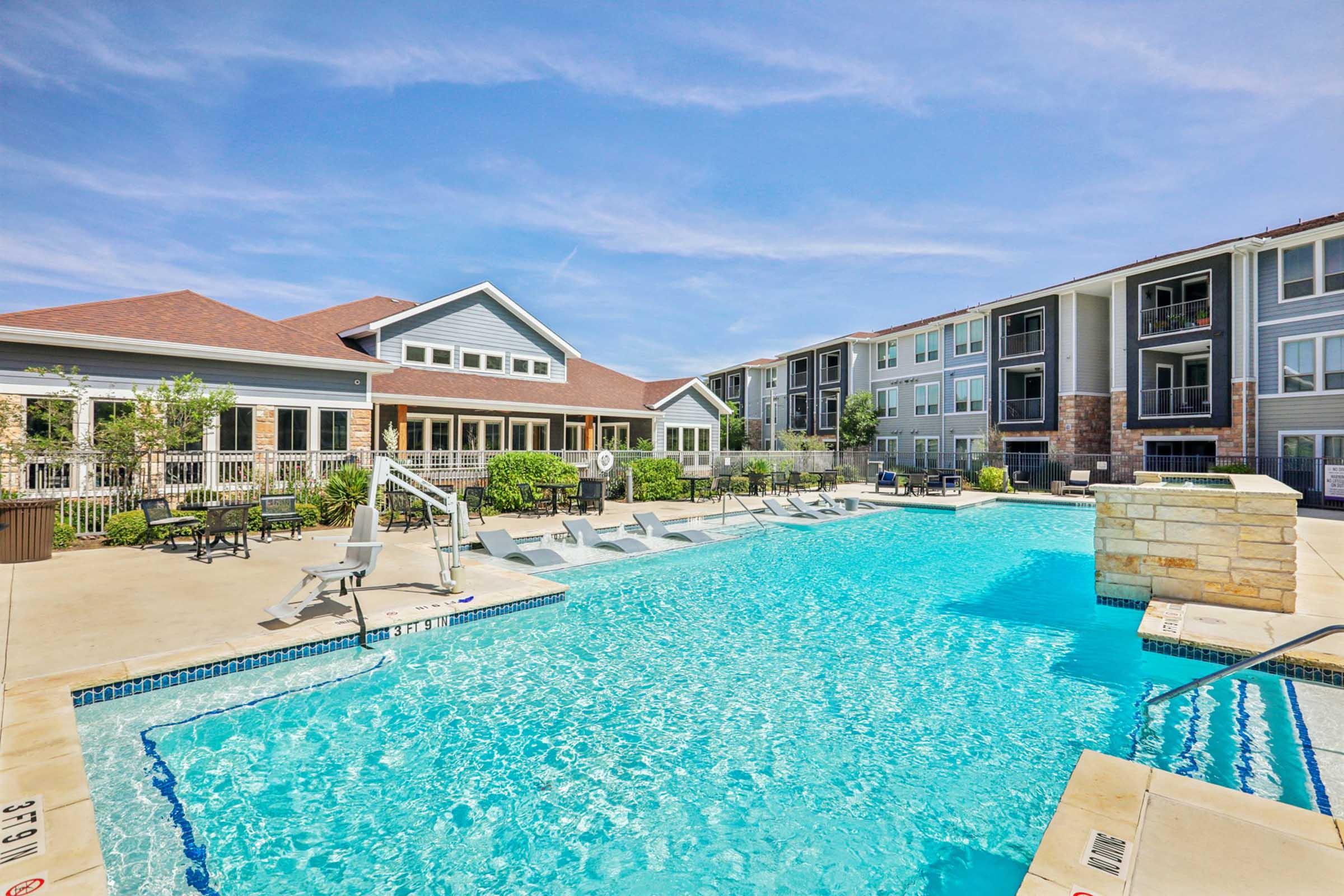A sparkling outdoor swimming pool surrounded by lounge chairs and greenery, with modern apartment buildings in the background. The area features sunny skies and a welcoming atmosphere, perfect for relaxation and leisure.