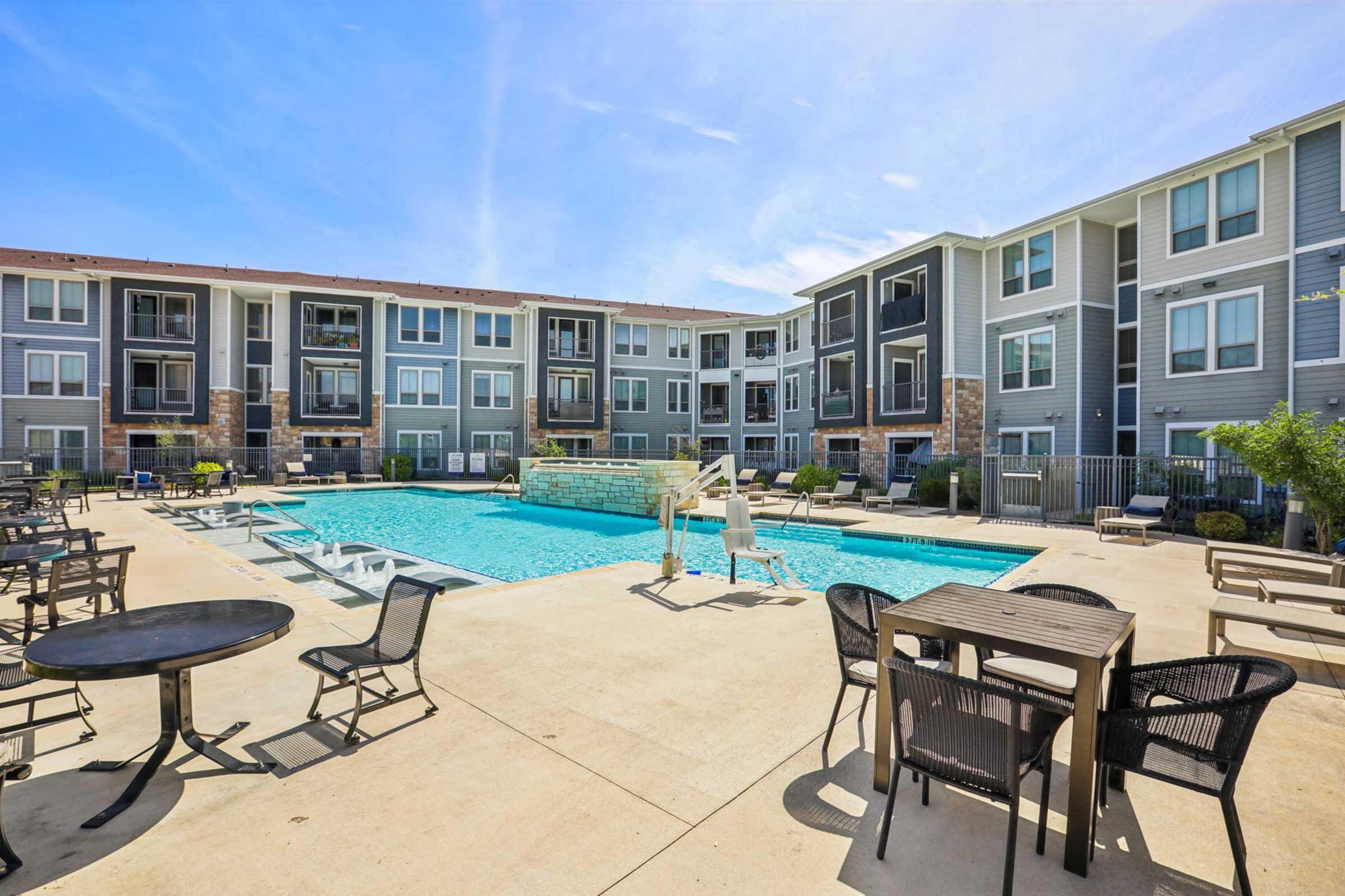 A well-maintained outdoor swimming pool surrounded by lounge chairs and tables, with modern apartment buildings in the background. The area is sunny and inviting, showcasing clear water and a landscaped environment, ideal for relaxation and social gatherings.