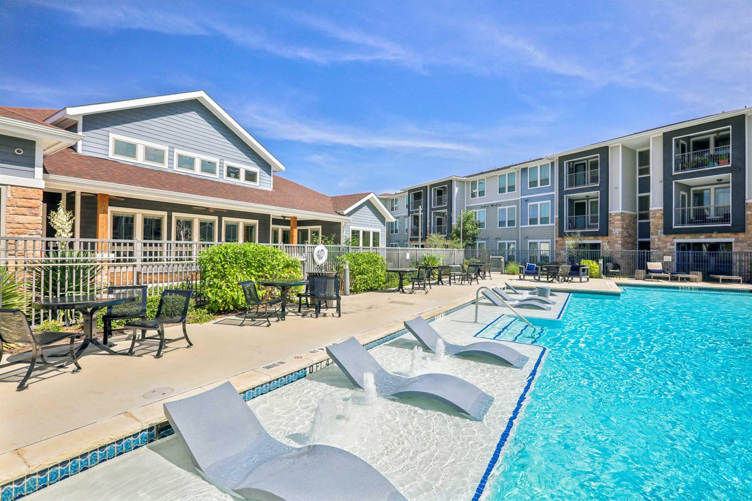 A sunny outdoor pool area with lounge chairs and a water feature. Surrounding the pool are trees and landscaped greenery, with two multi-story residential buildings in the background. The scene conveys a relaxing atmosphere, ideal for leisure and social gatherings.