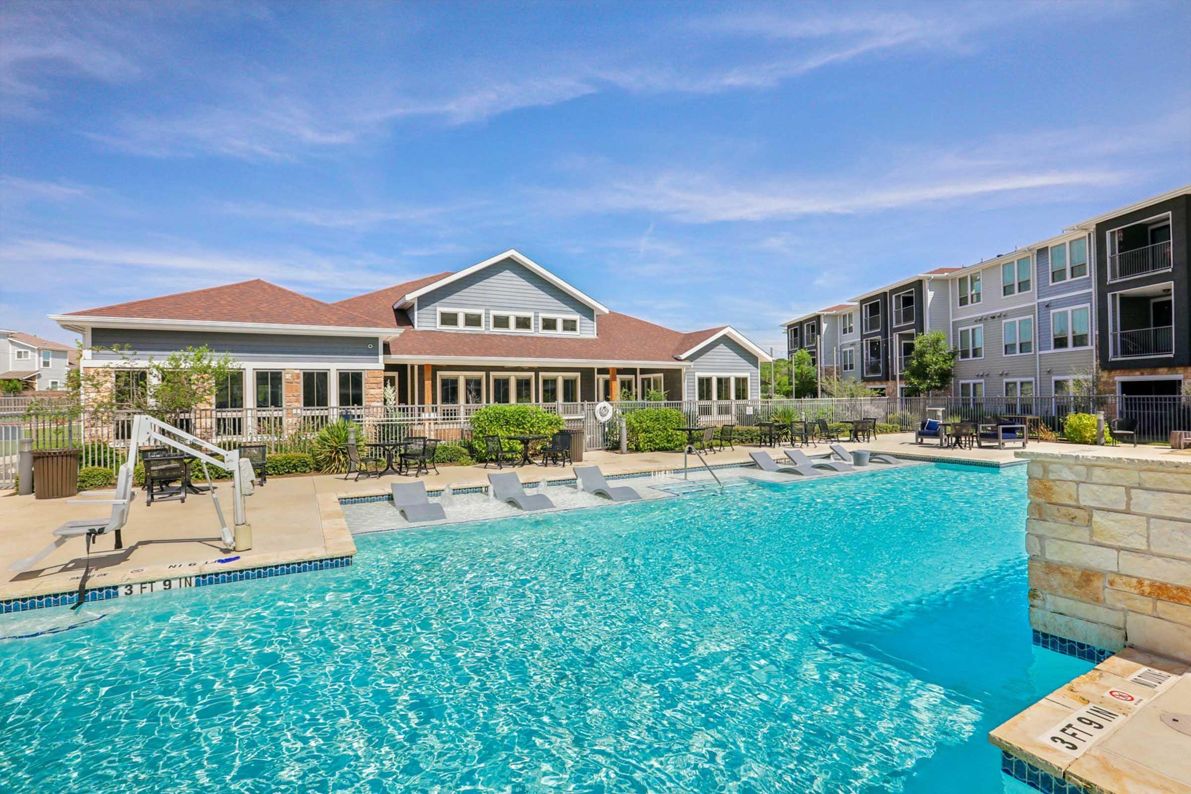 A refreshing outdoor swimming pool surrounded by lounge chairs and tables. In the background, a modern clubhouse with large windows can be seen, along with apartment buildings. The sky is clear with a few clouds, creating a bright and inviting atmosphere.