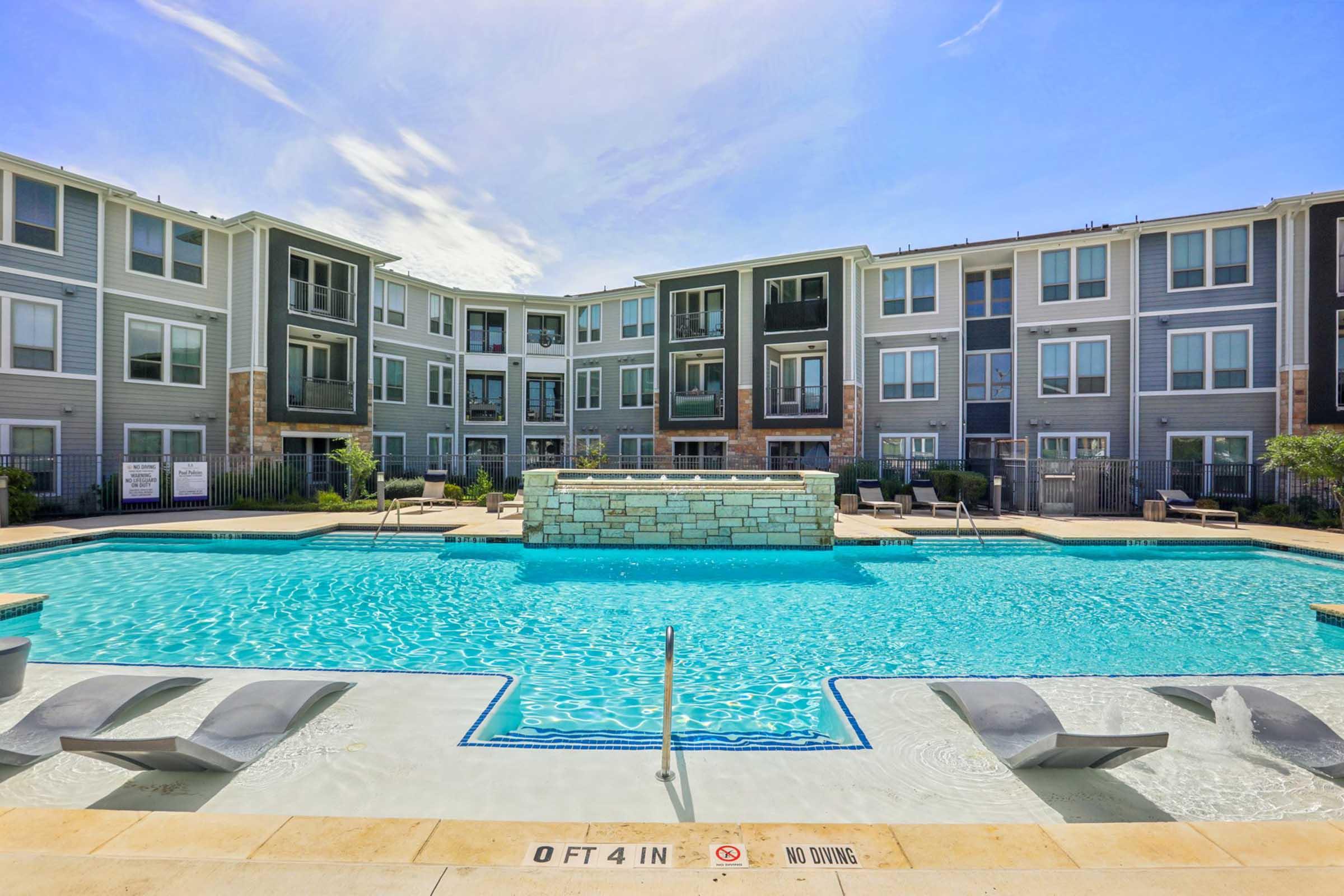 A spacious outdoor swimming pool surrounded by lounge chairs, with modern apartment buildings in the background. The pool features a stone fountain and clear blue water under a sunny sky. Safety signage is visible near the pool edge, and the overall setting is inviting and well-maintained.