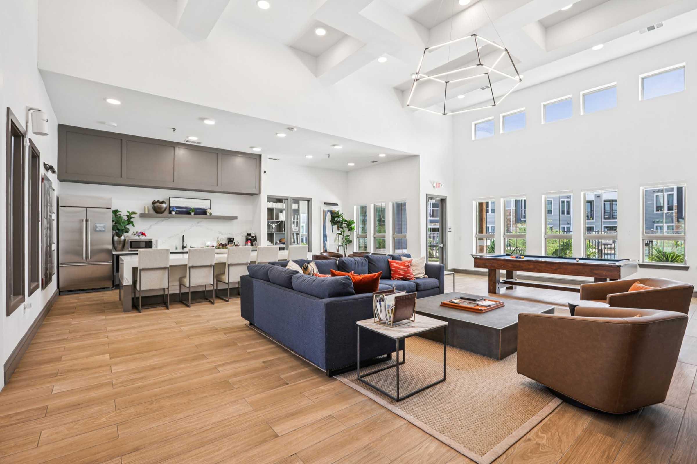 Interior of a modern living space featuring a bright, open layout. A cozy seating area with a dark blue sofa, orange accent pillows, and brown chairs surrounds a wooden coffee table. In the background, a kitchen area with stainless steel appliances and dining setup is visible. Large windows provide ample natural light.
