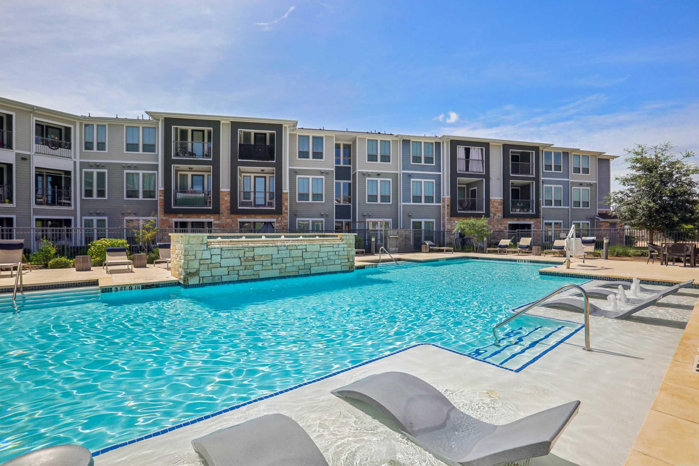 A spacious outdoor pool surrounded by modern apartment buildings. The pool features a stone wall and lounge chairs on the edge, with sunny blue skies in the background. Well-maintained landscaping adds to the inviting atmosphere of the area.