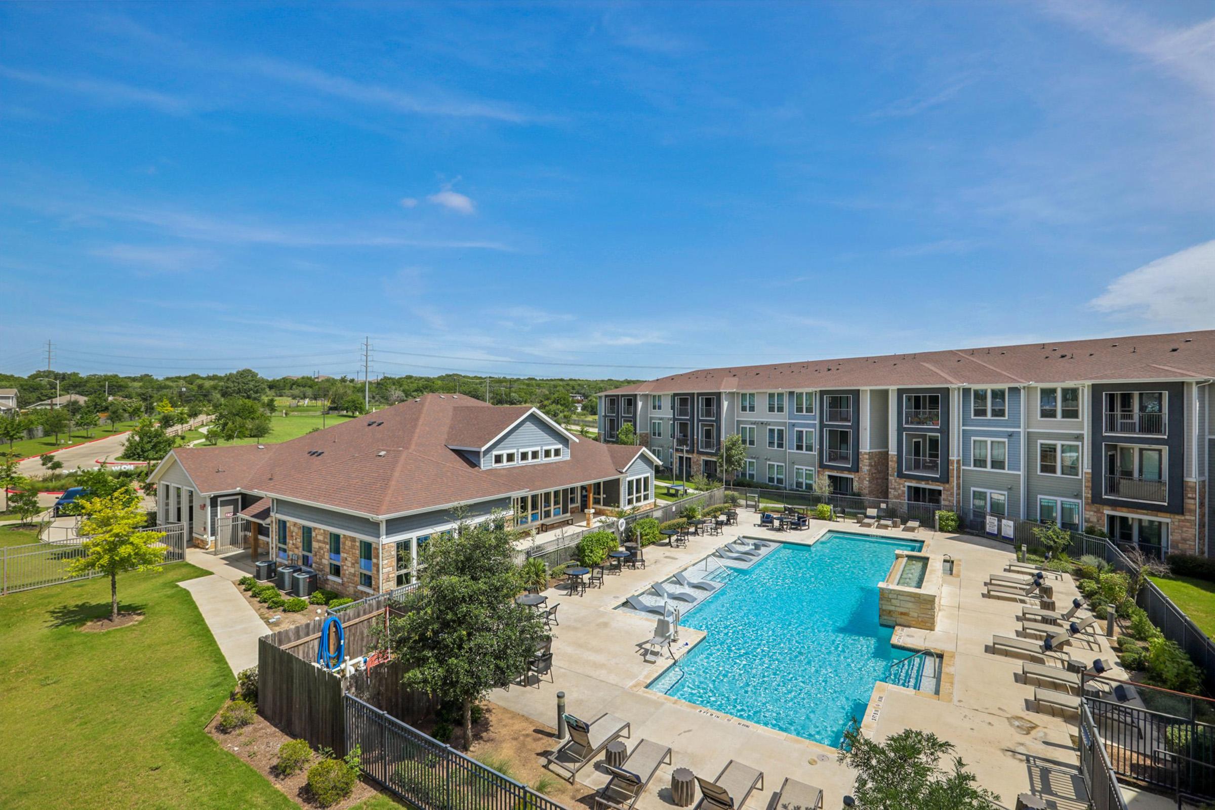 Aerial view of a modern apartment complex featuring a sparkling swimming pool surrounded by lounge chairs and greenery. The building has a mix of stone and siding exteriors, with a clear blue sky overhead. A well-maintained lawn and trees enhance the inviting atmosphere of the property.