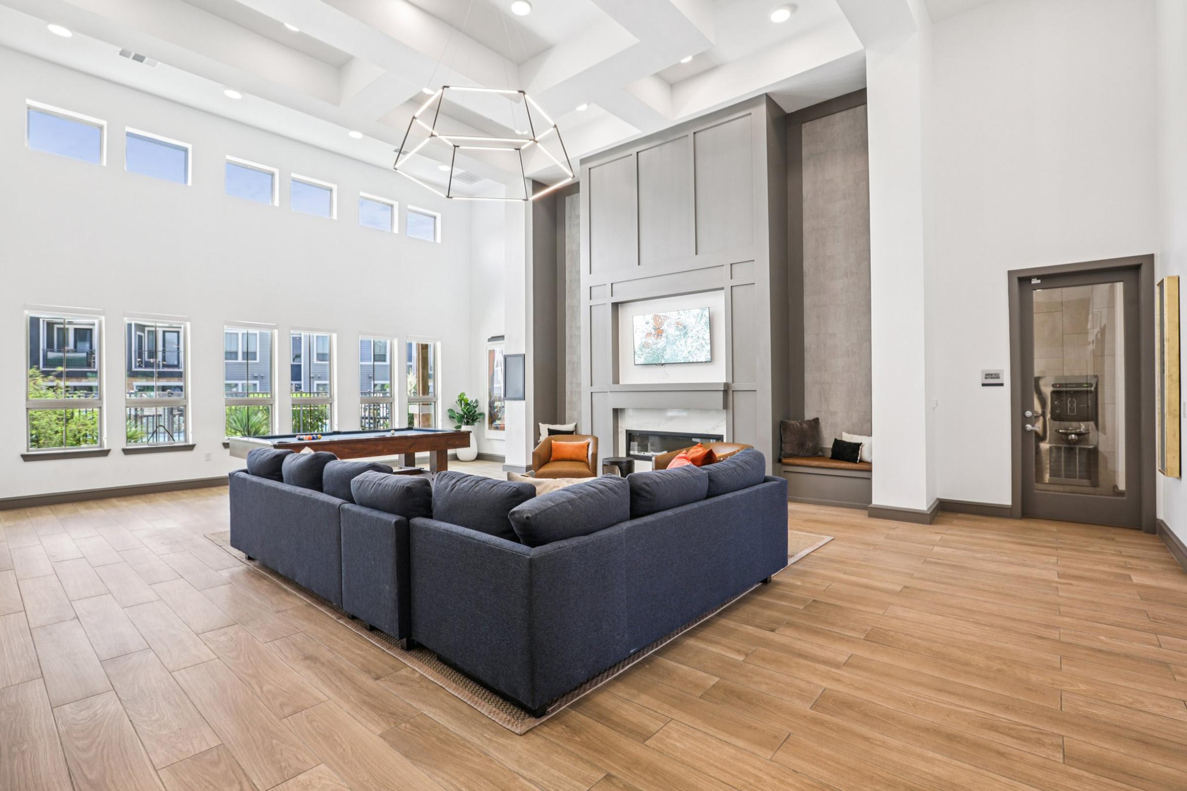 A spacious and modern living room featuring a large navy blue sectional sofa, a coffee table, and a fireplace. The room has tall windows allowing natural light to fill the space, complemented by high ceilings and a contemporary light fixture. There is a cozy nook with seating by the windows, enhancing the inviting atmosphere.