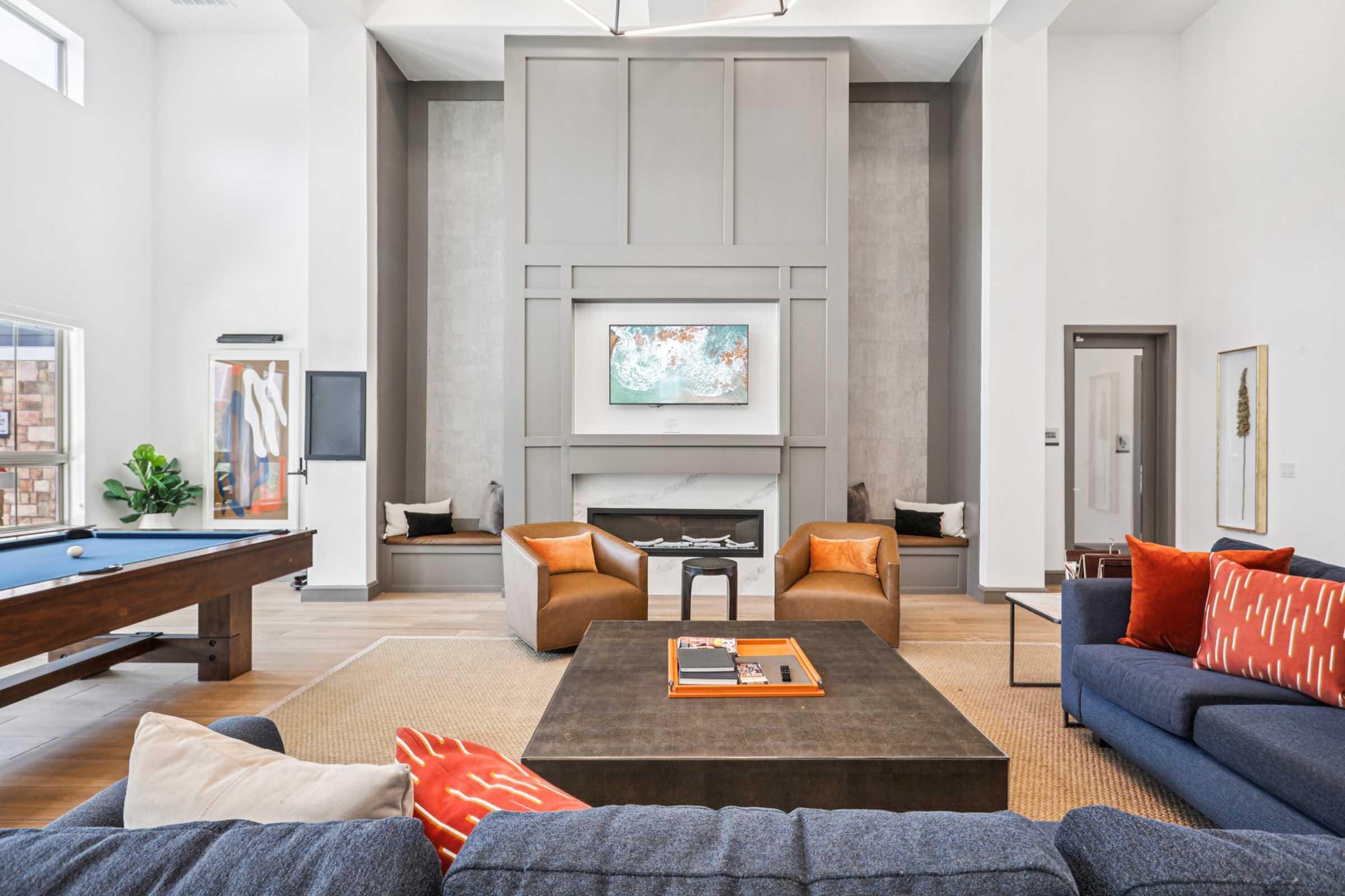 A modern living room featuring a high ceiling, large windows, and a neutral color palette. The space includes a dark coffee table, a plush sofa with vibrant orange and blue cushions, and two armchairs. A minimalist artwork hangs above a sleek fireplace, with a pool table in the background and decorative plants placed around the room.