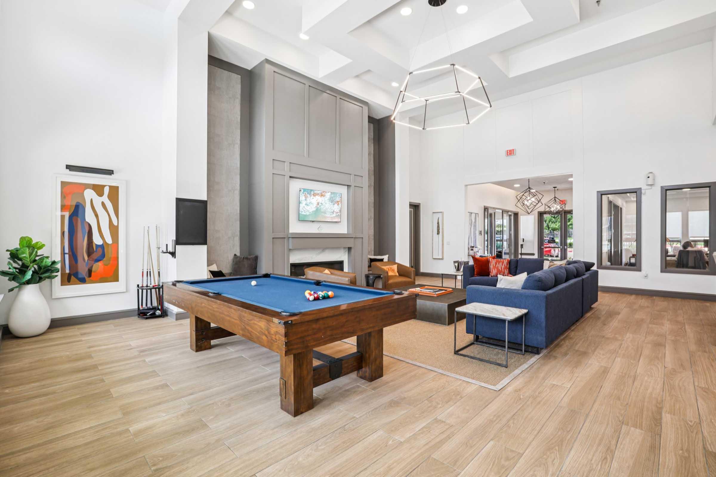 Interior of a modern communal living space featuring a pool table, comfortable seating area with a blue couch, large windows, and stylish decor. The space is well-lit with high ceilings and contemporary furnishings, creating a welcoming atmosphere for socializing and relaxation.