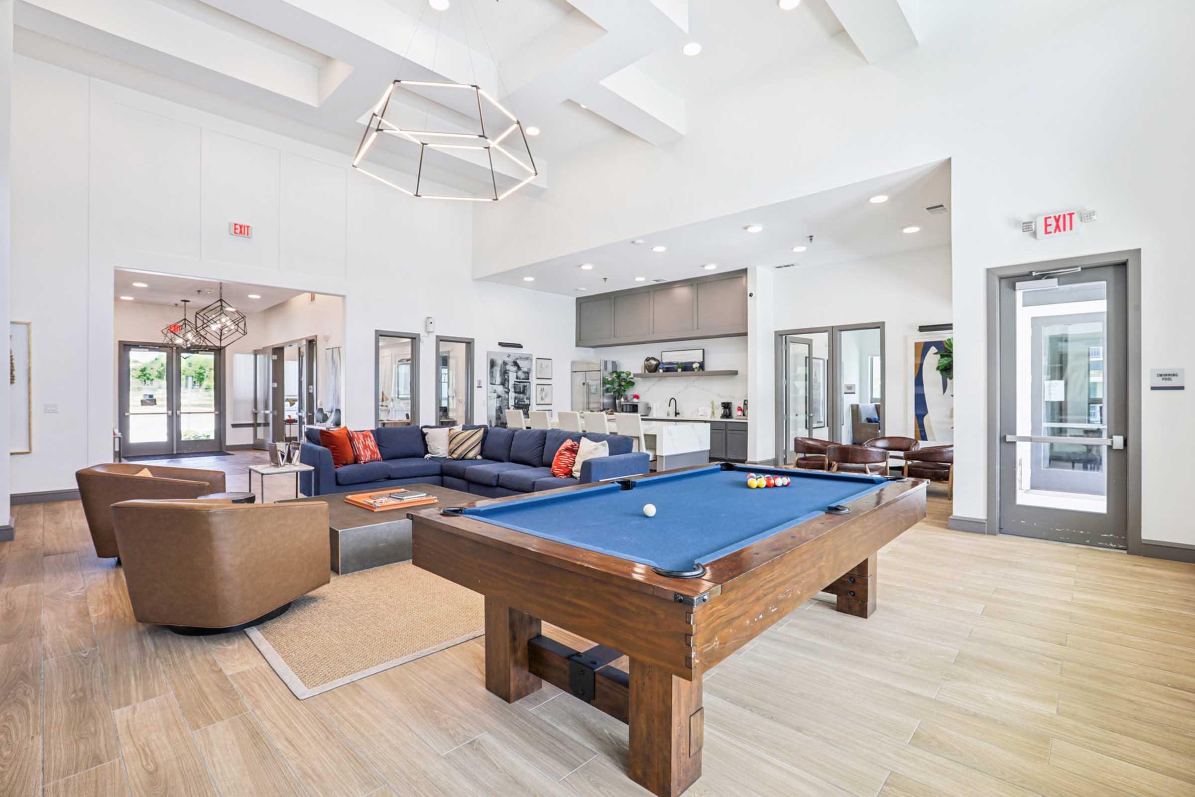 A modern lounge area featuring a pool table, a large sectional sofa with decorative pillows, and two brown armchairs. The room has large windows, stylish lighting fixtures, and a neutral color palette, creating a bright and inviting atmosphere. A kitchen area is visible in the background.