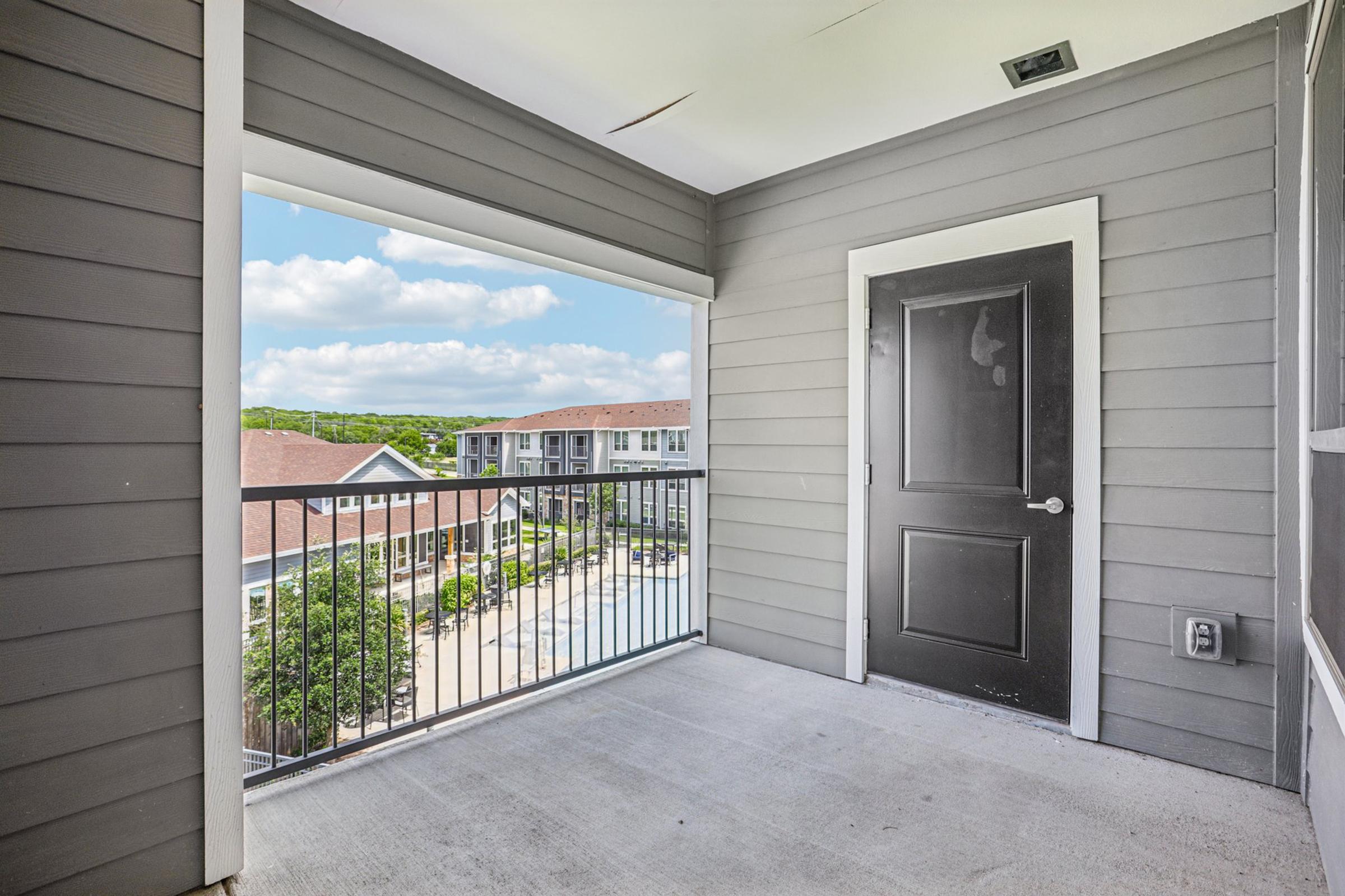 A balcony with a gray exterior, featuring a black door and a railing. The view includes a landscaped area with a pool and several buildings in the background against a blue sky with scattered clouds. The space is well-lit and appears to be part of an apartment complex.
