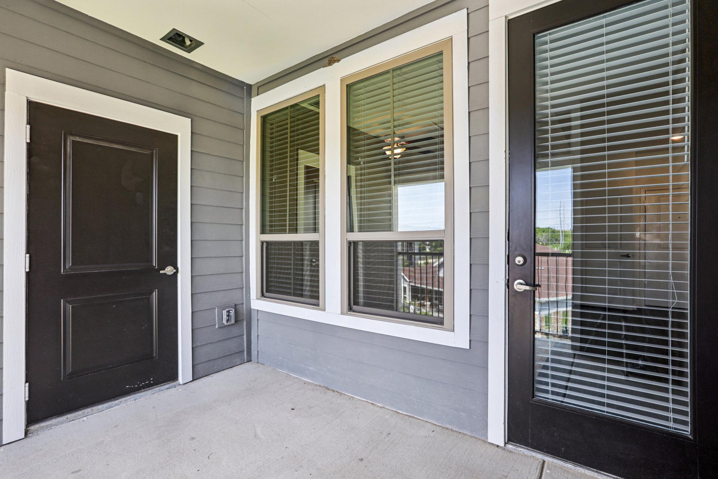 A modern entryway featuring a black front door on the left and a glass-paneled door on the right. The wall is clad in grey siding, and large windows allow natural light to illuminate the space. A ceiling fan is visible inside, adding to the contemporary feel of the entrance.