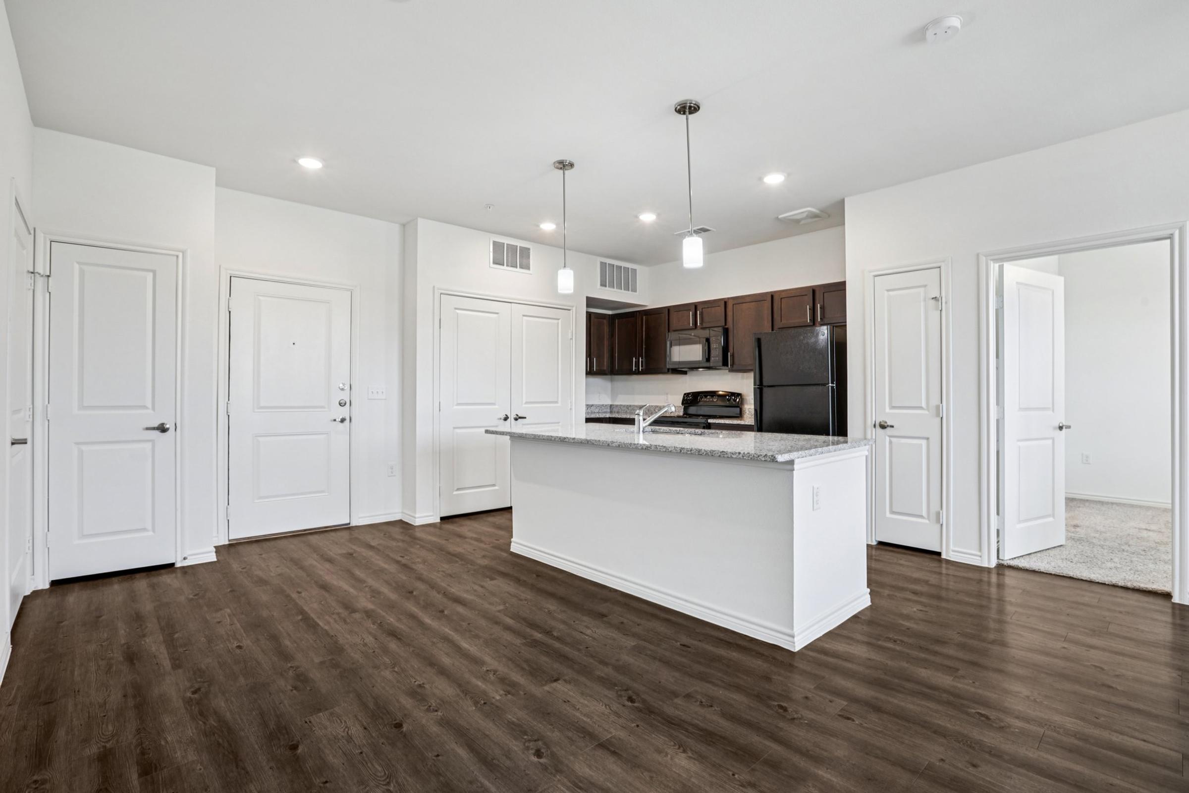 A modern, open-concept kitchen and living area featuring a kitchen island with a granite countertop, dark wood cabinetry, and stainless steel appliances. The space has light-colored walls, warm wood flooring, and bright overhead lighting, with doorways leading to other rooms.