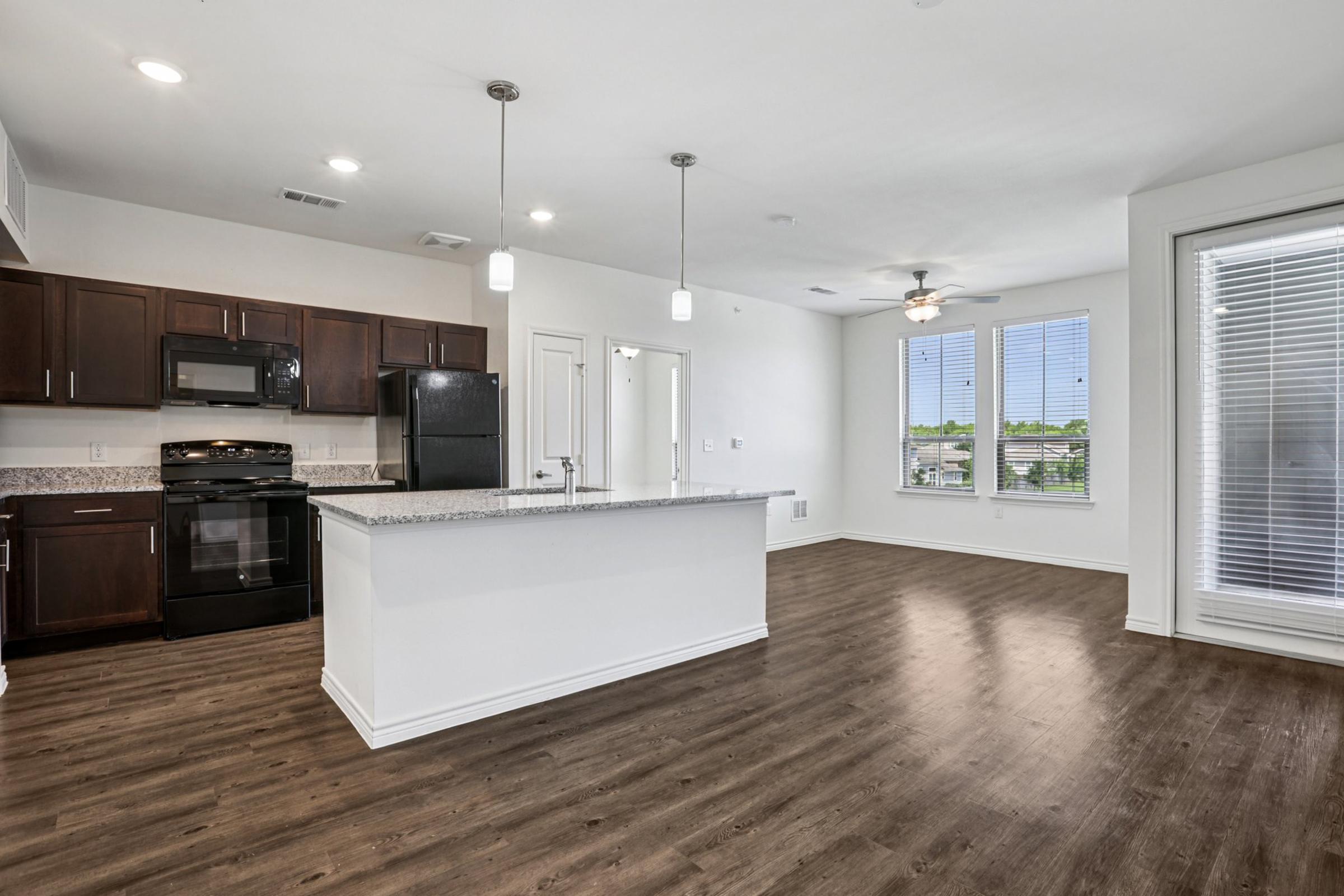 Modern open-concept kitchen and living area featuring dark wood cabinets, granite countertops, and stainless steel appliances. Bright natural light comes in through large windows, illuminating the spacious layout with wood-style flooring and contemporary light fixtures.