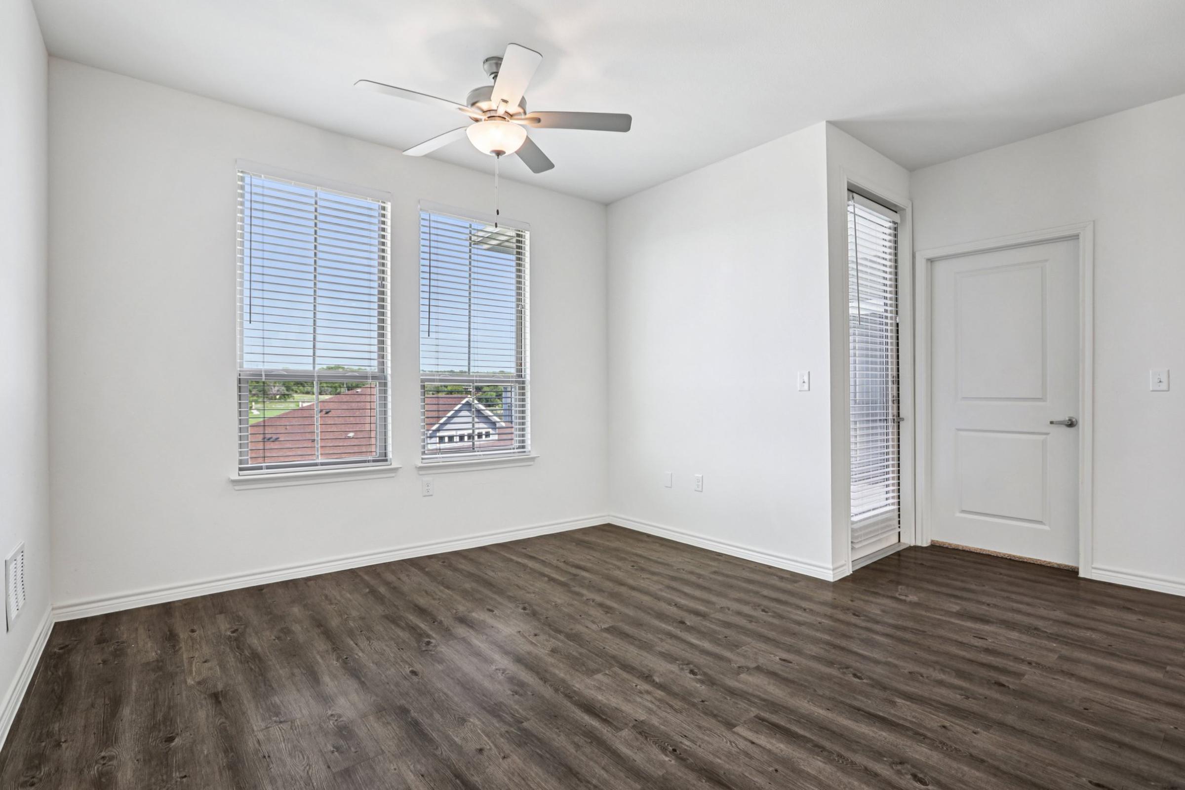A bright, empty room with large windows and blinds, a ceiling fan, and wood-like laminate flooring. The walls are painted white, and there is a door leading to another area. The view outside includes rooftops and green trees, creating a spacious and airy atmosphere.