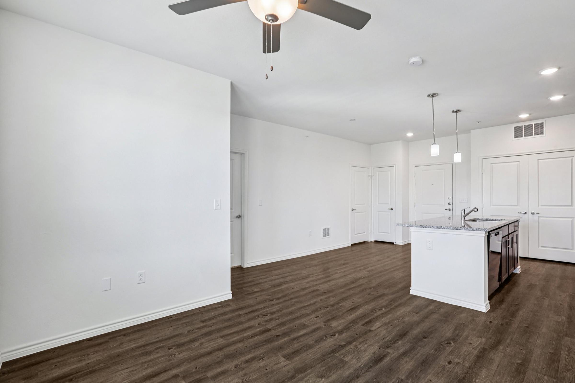 A modern, empty living area featuring light-colored walls and a ceiling fan. The space has hardwood floors and includes a kitchen area with a central island and pendant lighting. Two doors lead to additional rooms, and there are windows allowing natural light to enter.