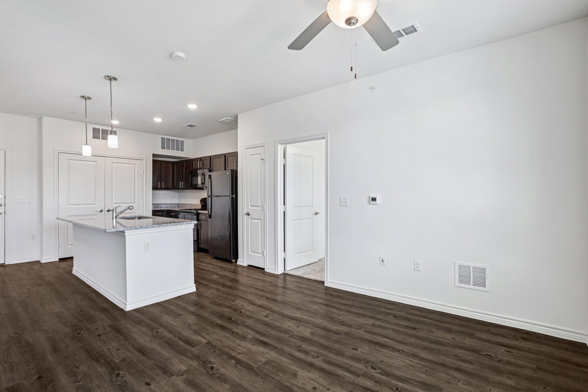 Modern open-concept living space featuring a kitchen island with bar seating, stainless steel appliances, and a ceiling fan. Light-colored walls and wooden flooring create a bright atmosphere. A doorway leads to a separate room, while natural light enhances the overall warmth of the area.