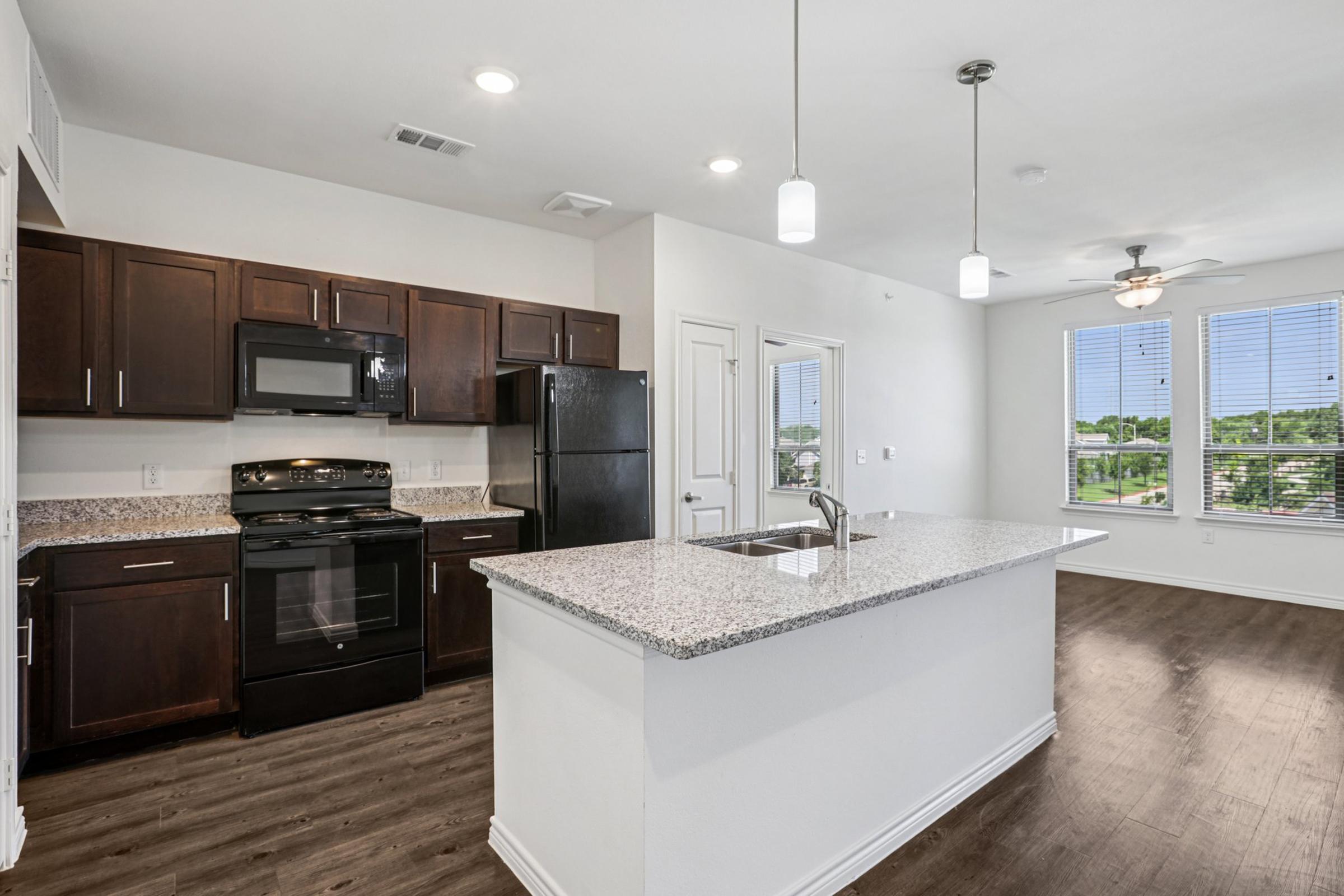 Modern kitchen featuring dark wooden cabinets, a black refrigerator, and a black stove. There is a large granite countertop with a sink and pendant lighting above. The open layout connects to a bright living space with large windows, allowing natural light to fill the room.