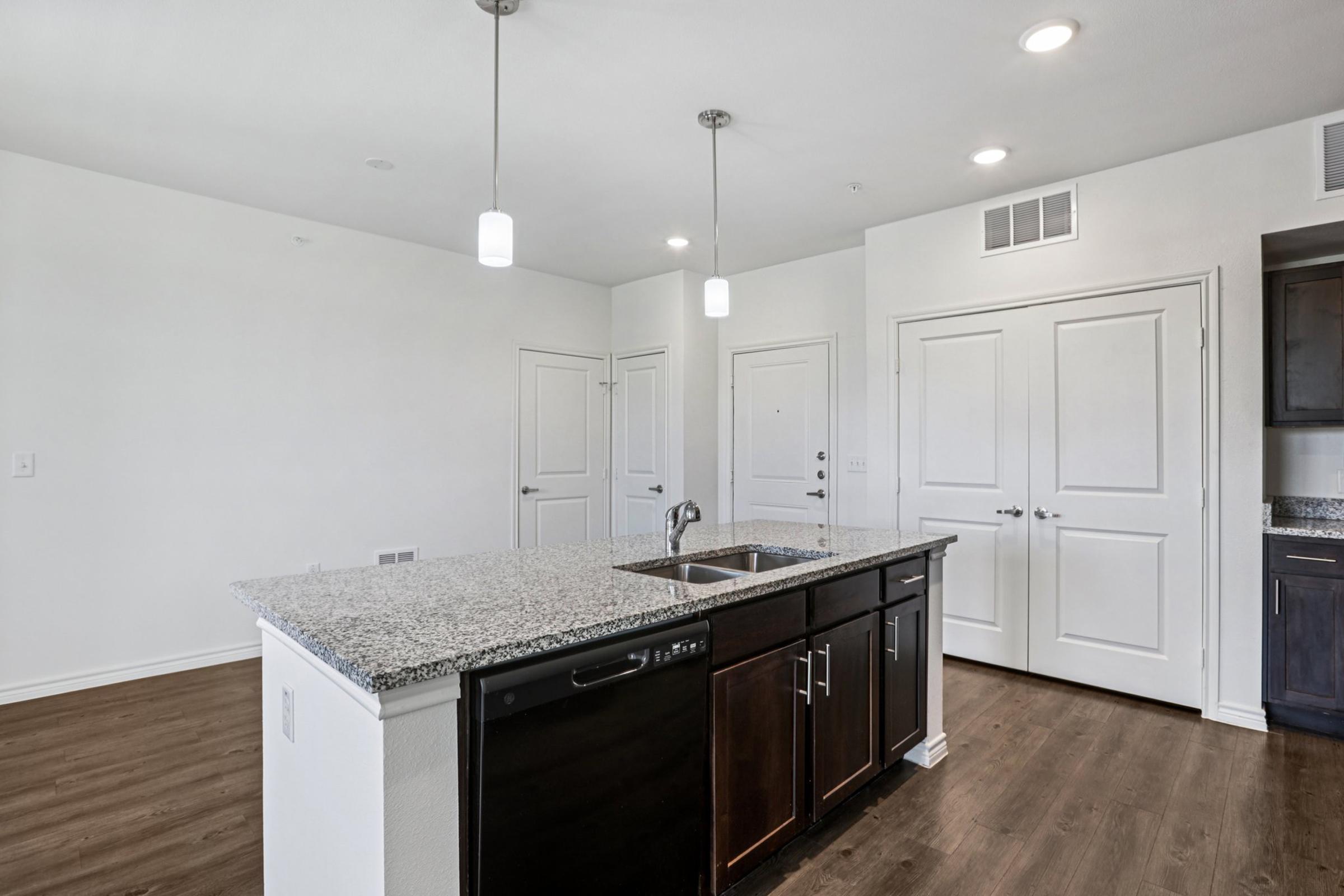 Modern kitchen featuring a central island with a sink, dark wooden cabinetry, and white walls. The space is well-lit with three pendant lights hanging from the ceiling. A hardwood floor complements the kitchen design, and there are two doors leading to adjacent rooms.