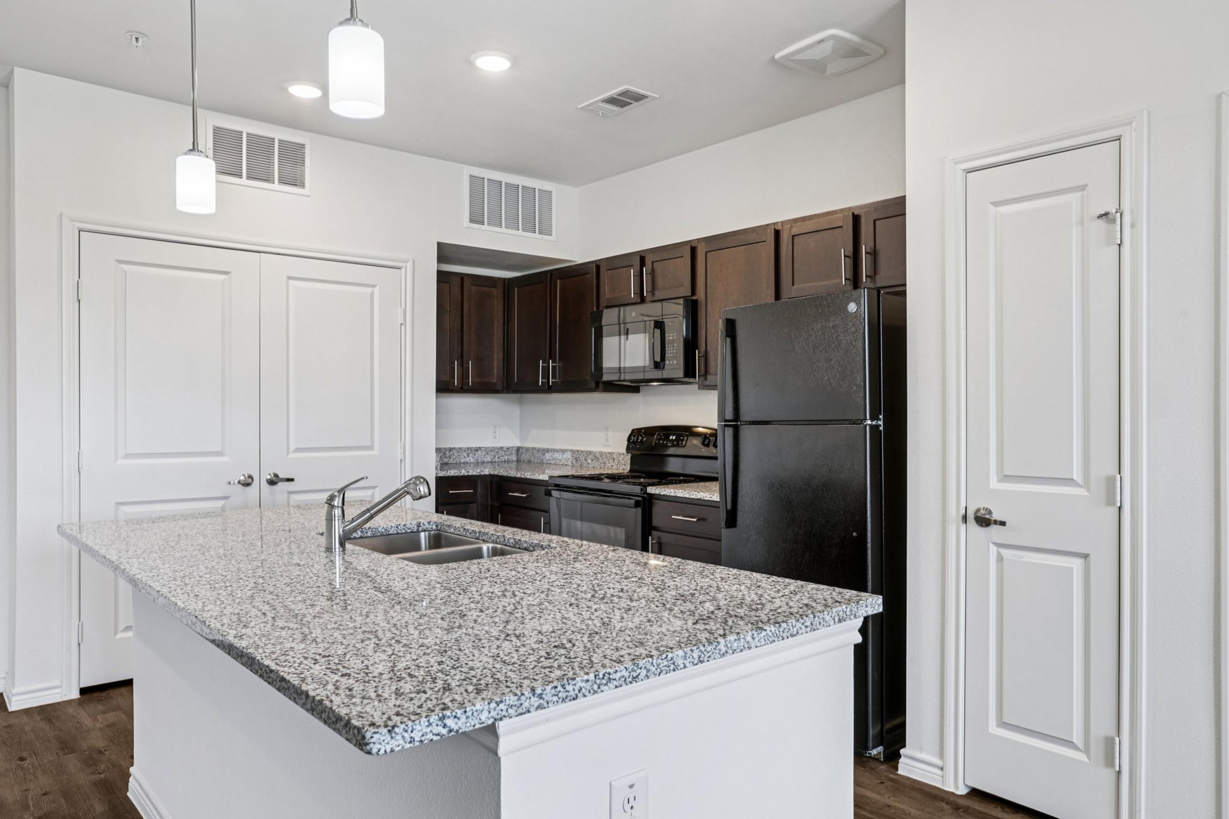 Modern kitchen interior featuring dark wood cabinets, a granite countertop, stainless steel appliances including a microwave and an oven, a black refrigerator, and pendant lighting above the island. The room has a light-colored wall and wooden flooring, creating a sleek and contemporary atmosphere.