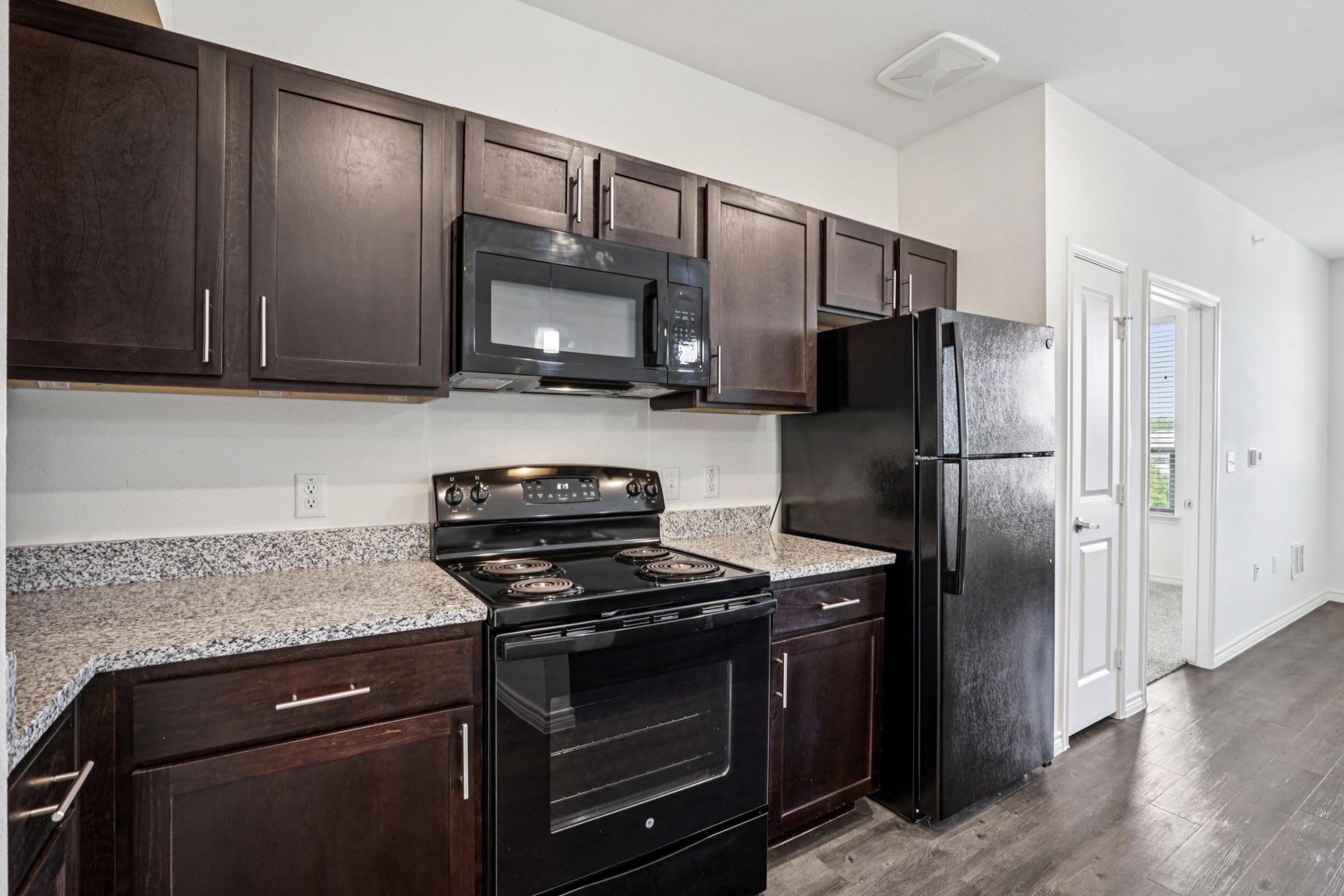 Modern kitchen featuring dark wooden cabinets, a granite countertop, black appliances including an oven, microwave, and refrigerator. The space is well-lit with natural light and has a clean, contemporary design. The flooring is a dark wood, complementing the overall aesthetic.