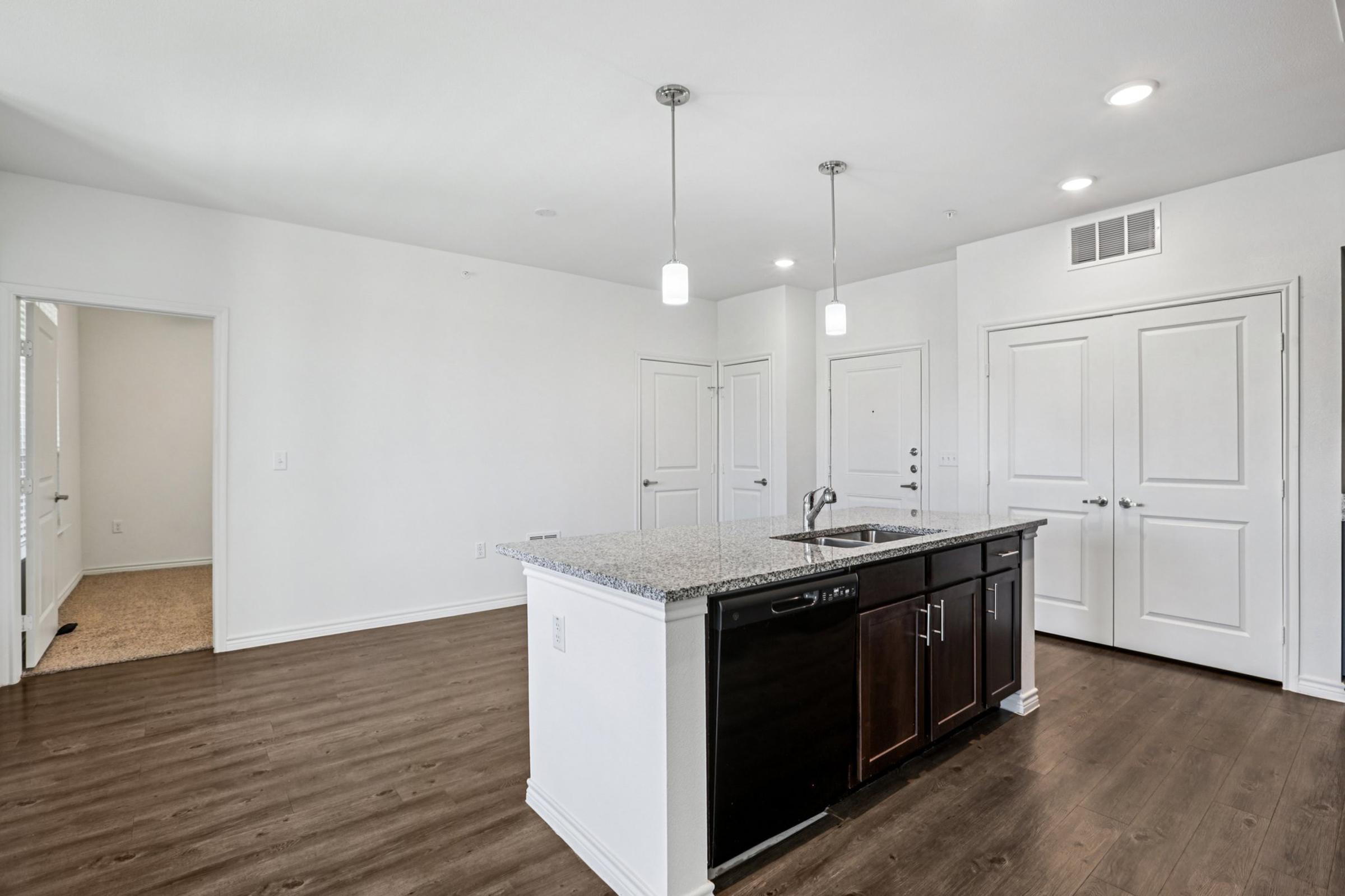 A modern kitchen with an island featuring a granite countertop, black appliances, and pendant lighting. The space has light-colored walls and wooden flooring, with an open layout leading to a living area and a doorway. Bright natural light filters through the windows, creating a spacious feel.