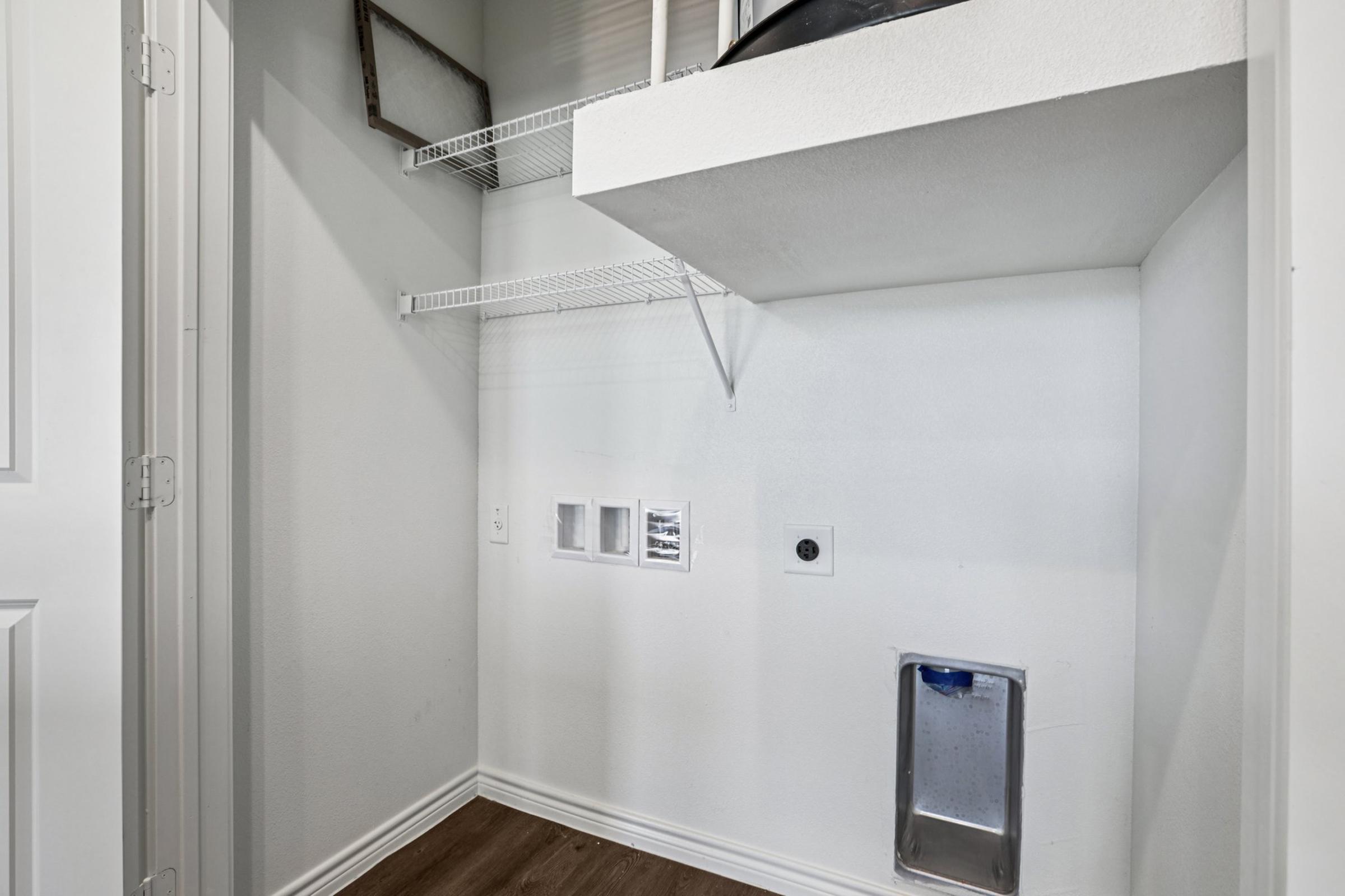 A small, empty laundry room with white walls and a wooden floor. There are shelves along one wall, with a few hooks for hanging items. The lower part features a wall-mounted pet door and electrical outlets. The space appears clean and organized, suitable for laundry tasks or storage.