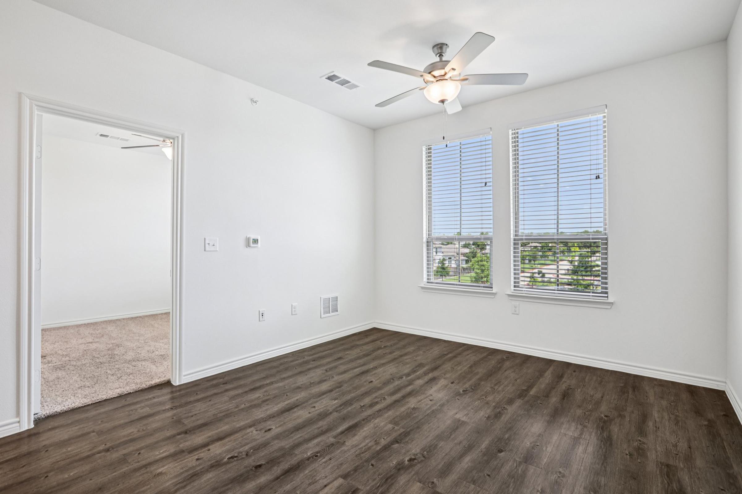 A well-lit, empty room featuring light wood flooring, two large windows with blinds, and a ceiling fan. The room has plain white walls and a door opening to another area. The overall ambiance is clean and modern, ideal for various decorating possibilities.