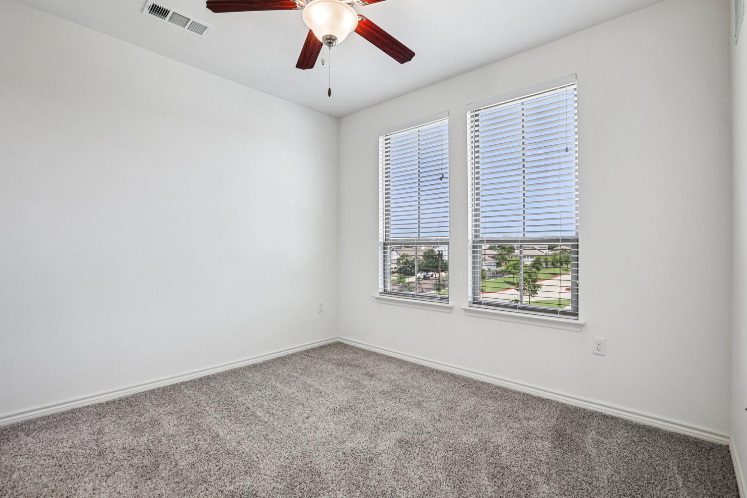 Empty room with beige carpet flooring, two windows with white blinds, and a ceiling fan with wooden blades. The walls are painted white, and natural light is streaming in through the windows, offering a view of the outside.