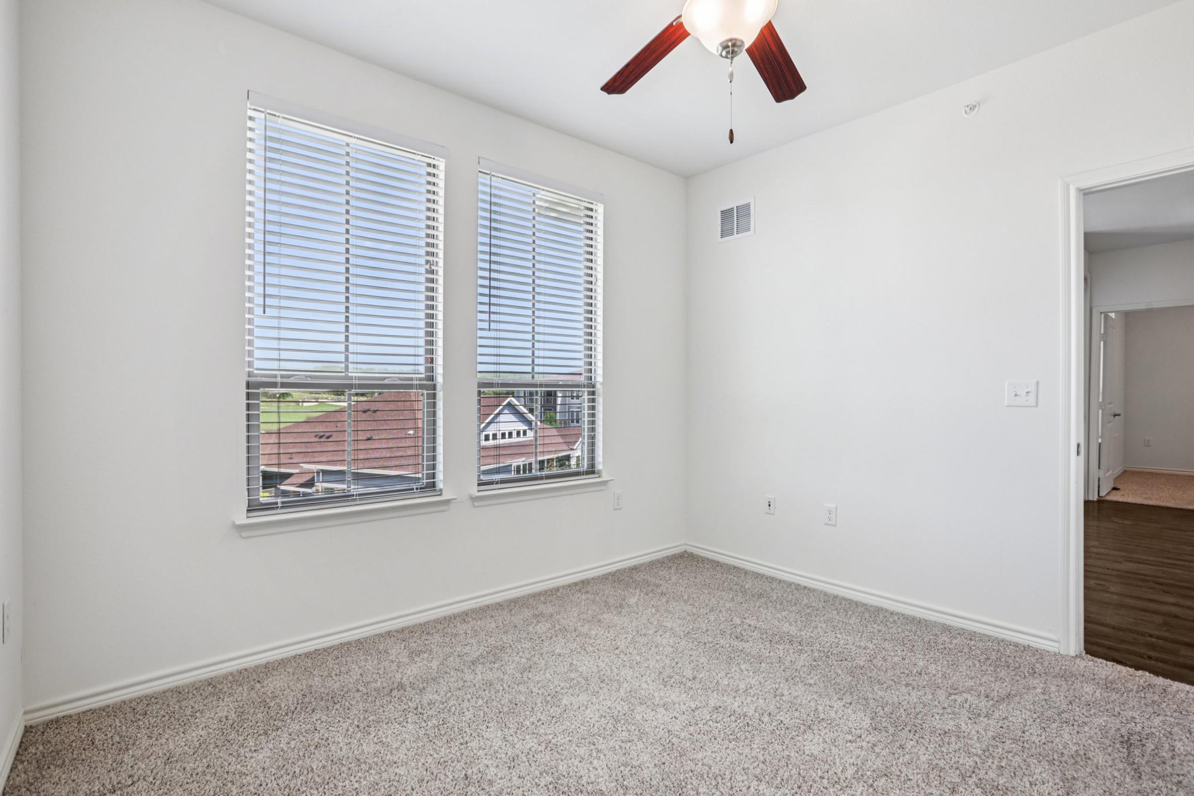 A bright, empty room featuring white walls and two large windows with blinds. Natural light floods the space, highlighting the beige carpet flooring. In the background, there is a glimpse of a neighboring building with a roof. The room also has a ceiling fan and an open doorway leading to another area.