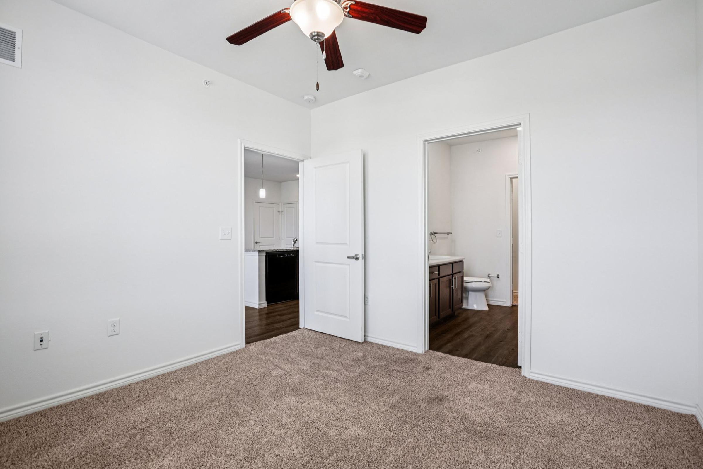 Room with beige carpet and white walls, featuring a ceiling fan with dark wood blades. A door leads to a bathroom visible in the background, while another door opens to an adjacent room. The space is well-lit and uncluttered, creating a neat and modern appearance.