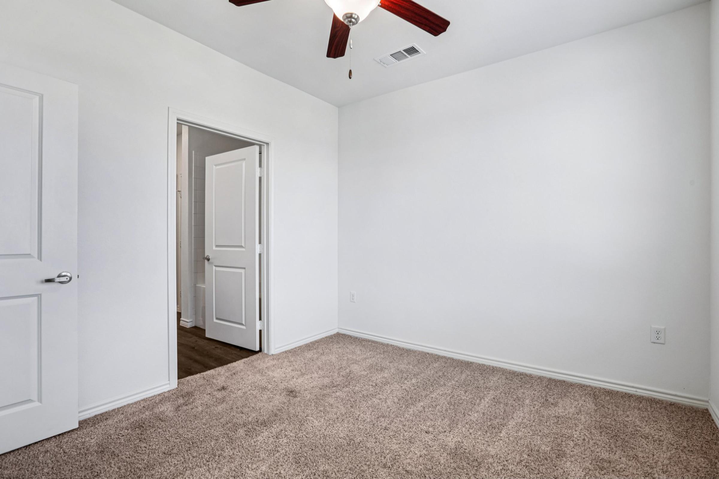 A spacious, empty room with light gray walls and plush beige carpet. A white ceiling features a ceiling fan, and there is a closed white door on the left leading to another room. A doorway in the back suggests an entrance to a bathroom or another area. The overall atmosphere is bright and uncluttered.