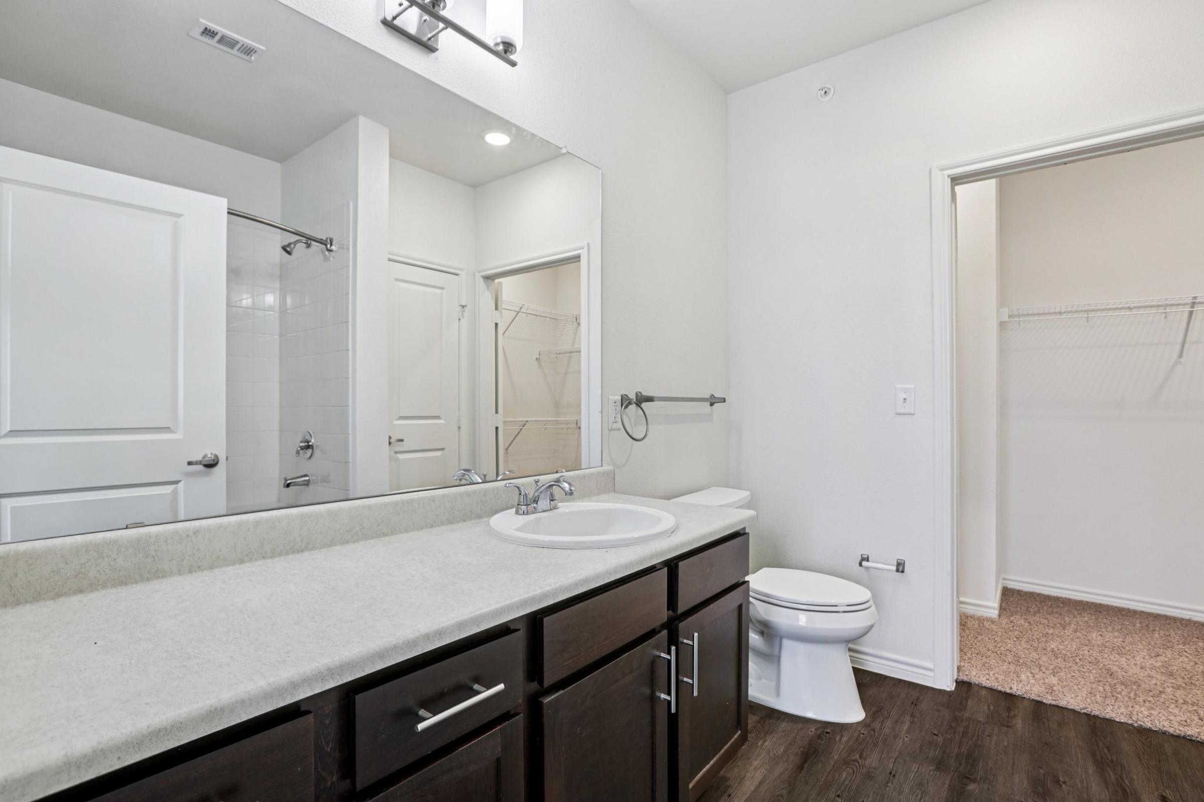 A modern bathroom featuring a large vanity with a sink and dark wood cabinets, a white toilet, and a walk-in closet. The walls are light-colored, and there's a shower area visible with a glass door. The floor has a wooden appearance, adding warmth to the space.