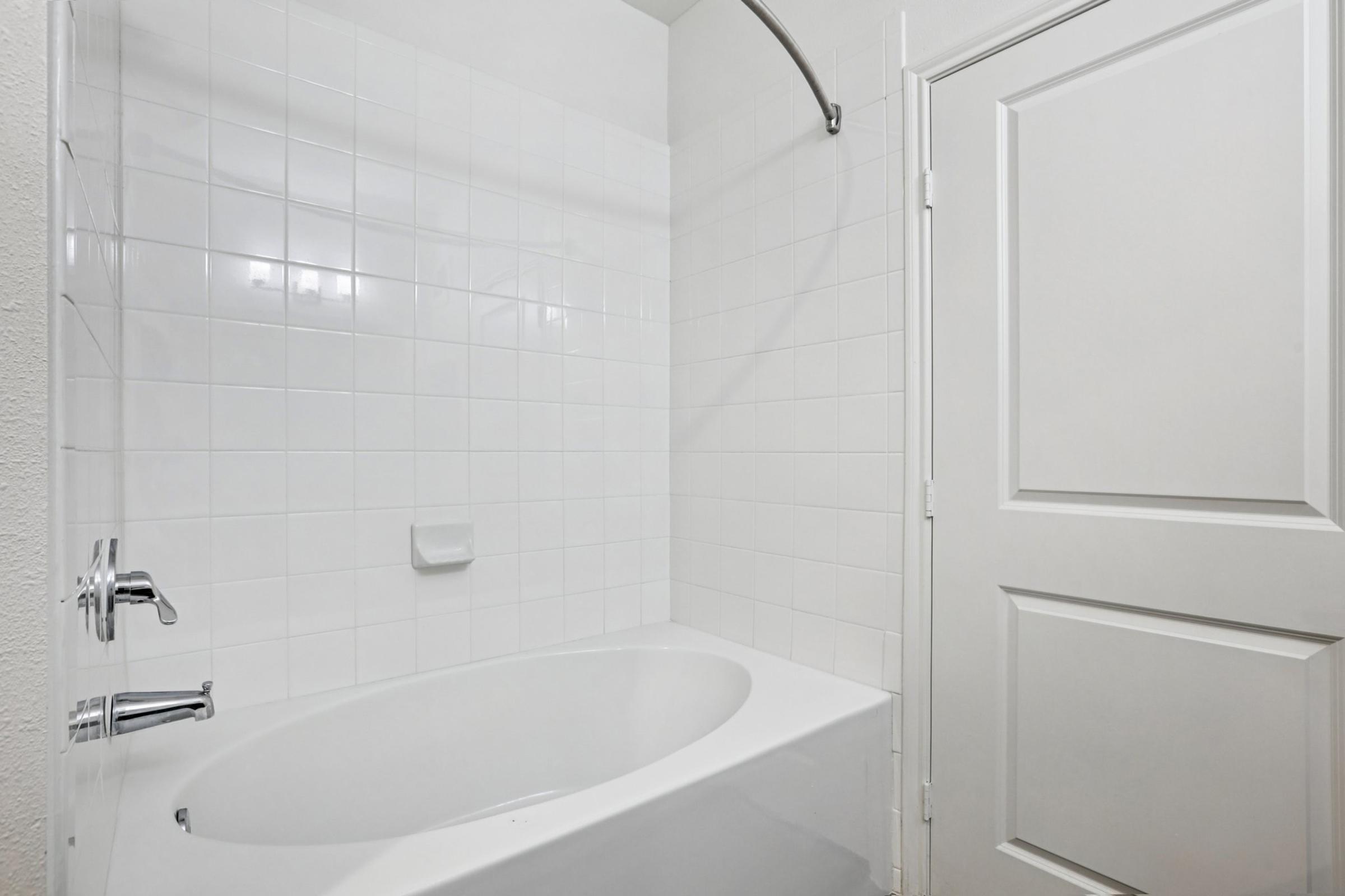Bathtub with a curved shower rod and white tile walls in a clean, modern bathroom. The door is closed, and a soap dish is mounted on the wall. The overall aesthetic is minimalistic and bright, featuring neutral colors.