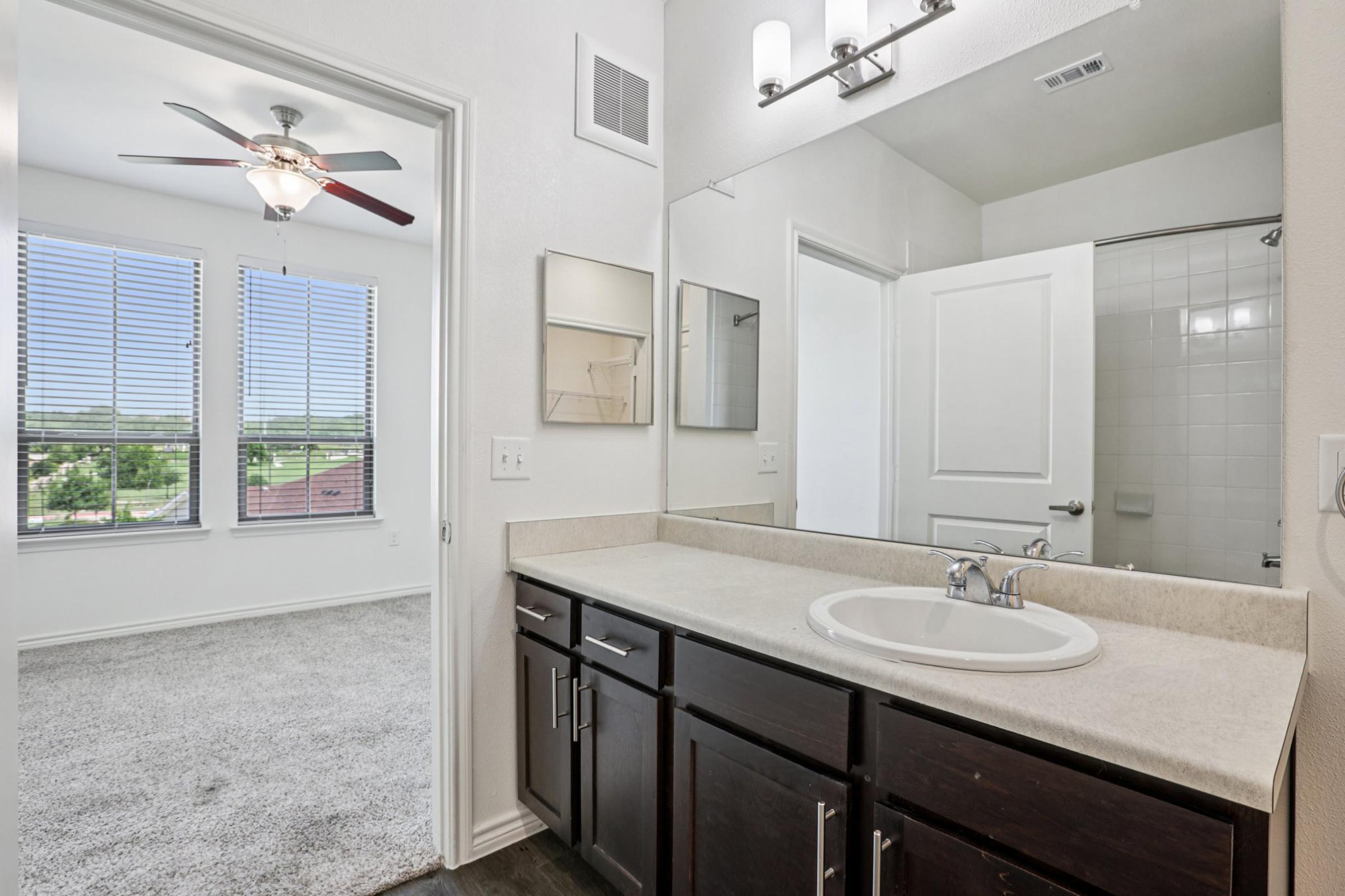 A modern bathroom featuring a double sink vanity with dark wood cabinetry, a large mirror, and a shower area. A bright bedroom with large windows and a ceiling fan is visible through an open door, showcasing a carpeted floor and a view of the outdoors. The overall aesthetic is clean and contemporary.
