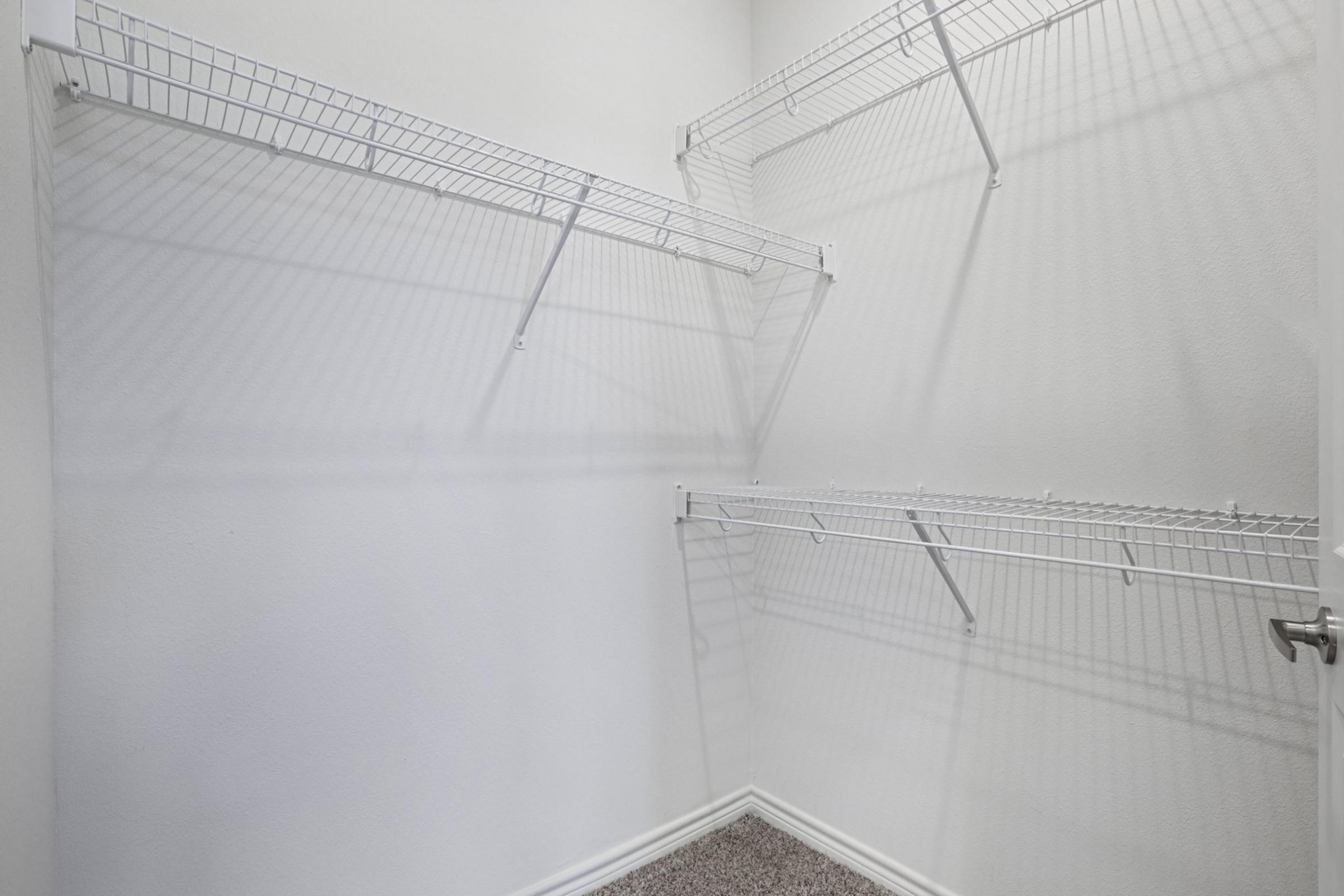 Empty closet with white wire shelving arranged on the walls. The walls are painted in a neutral light color, and the floor is covered in a soft carpet. The closet appears spacious and well-lit, providing ample storage options.