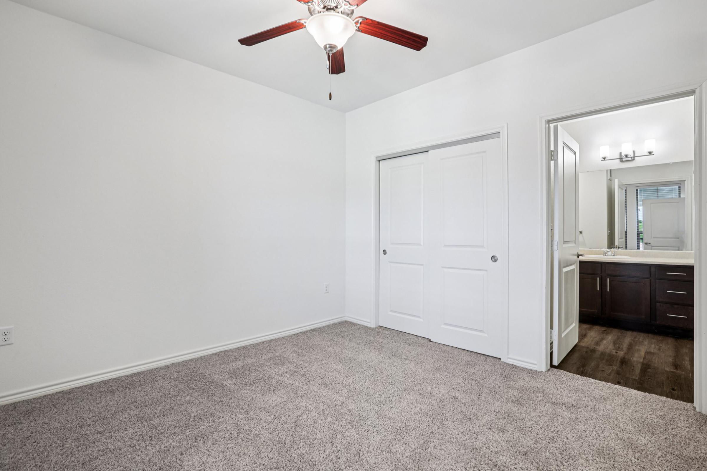 A well-lit room with a ceiling fan, featuring light-colored walls and carpeted flooring. To the right, there is a doorway leading to a bathroom with modern fixtures and a dark wood vanity. A closet with double doors is visible on the left. The overall atmosphere is clean and inviting.